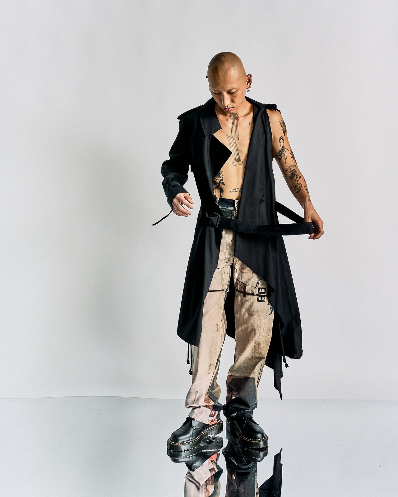 Man with a shaved head wearing an open black Maison Margiela trench coat, revealing tattoos on his chest and arms, paired with beige pants and platform shoes.