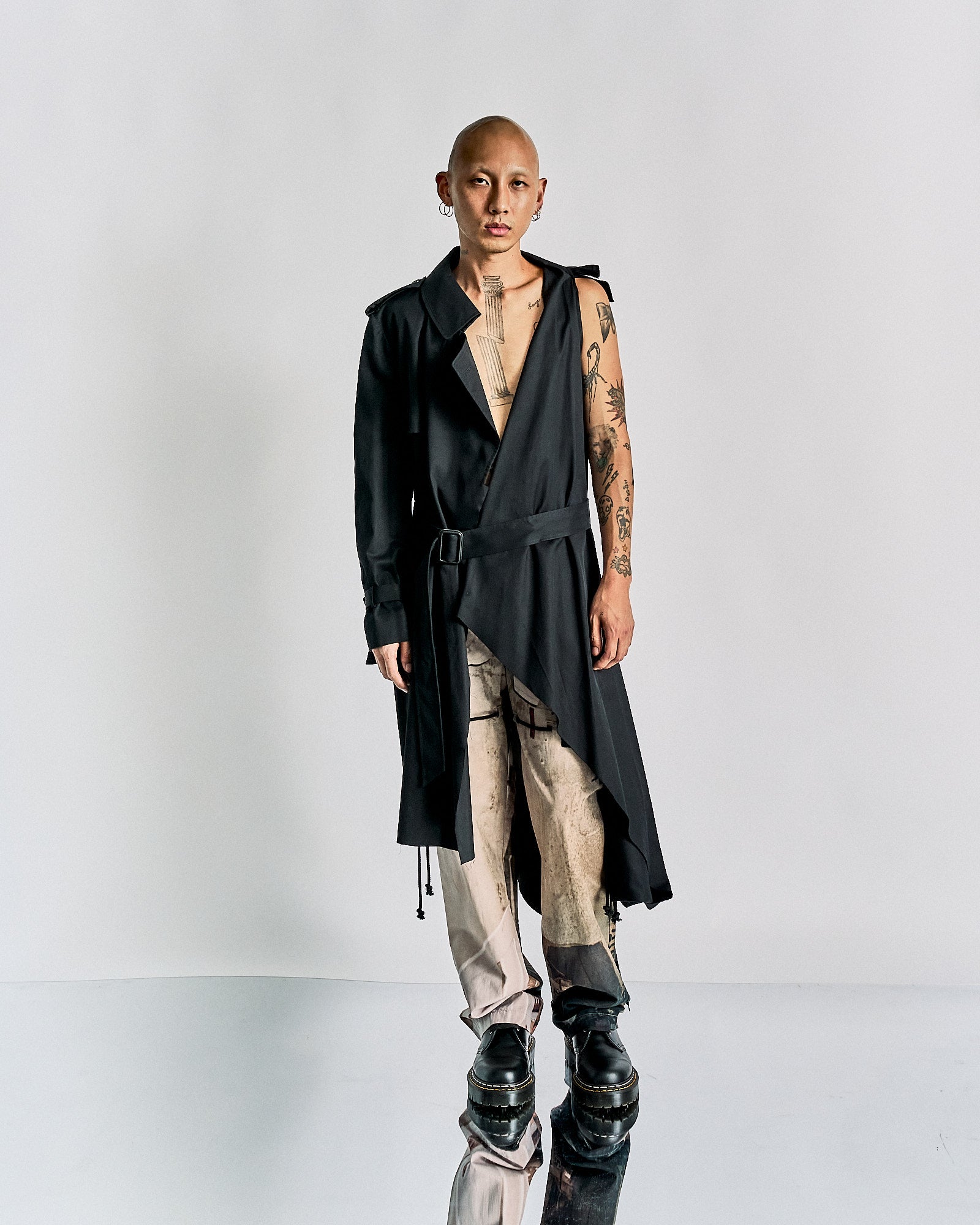 A person stands against a neutral background wearing an asymmetrical, black Maison Margiela wrap coat and light, patterned pants with black shoes