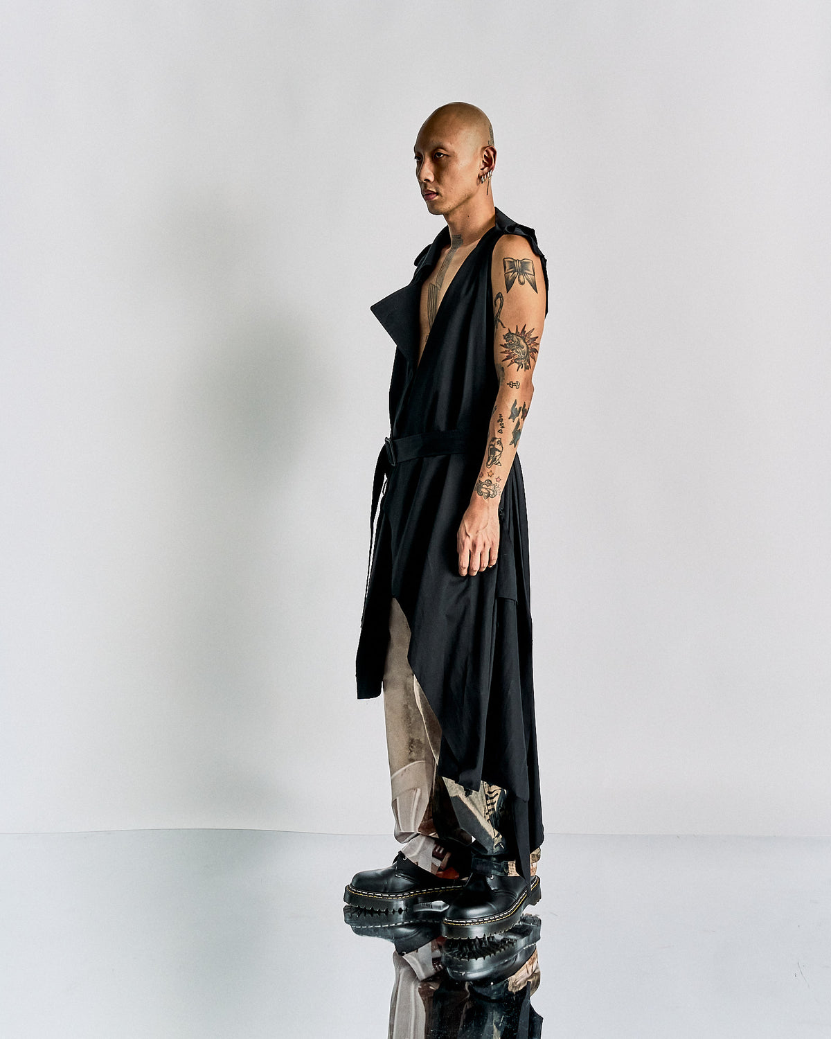 Man with a shaved head wearing an open black Maison Margiela trench coat, revealing tattoos on his chest and arms, paired with beige pants and platform shoes.