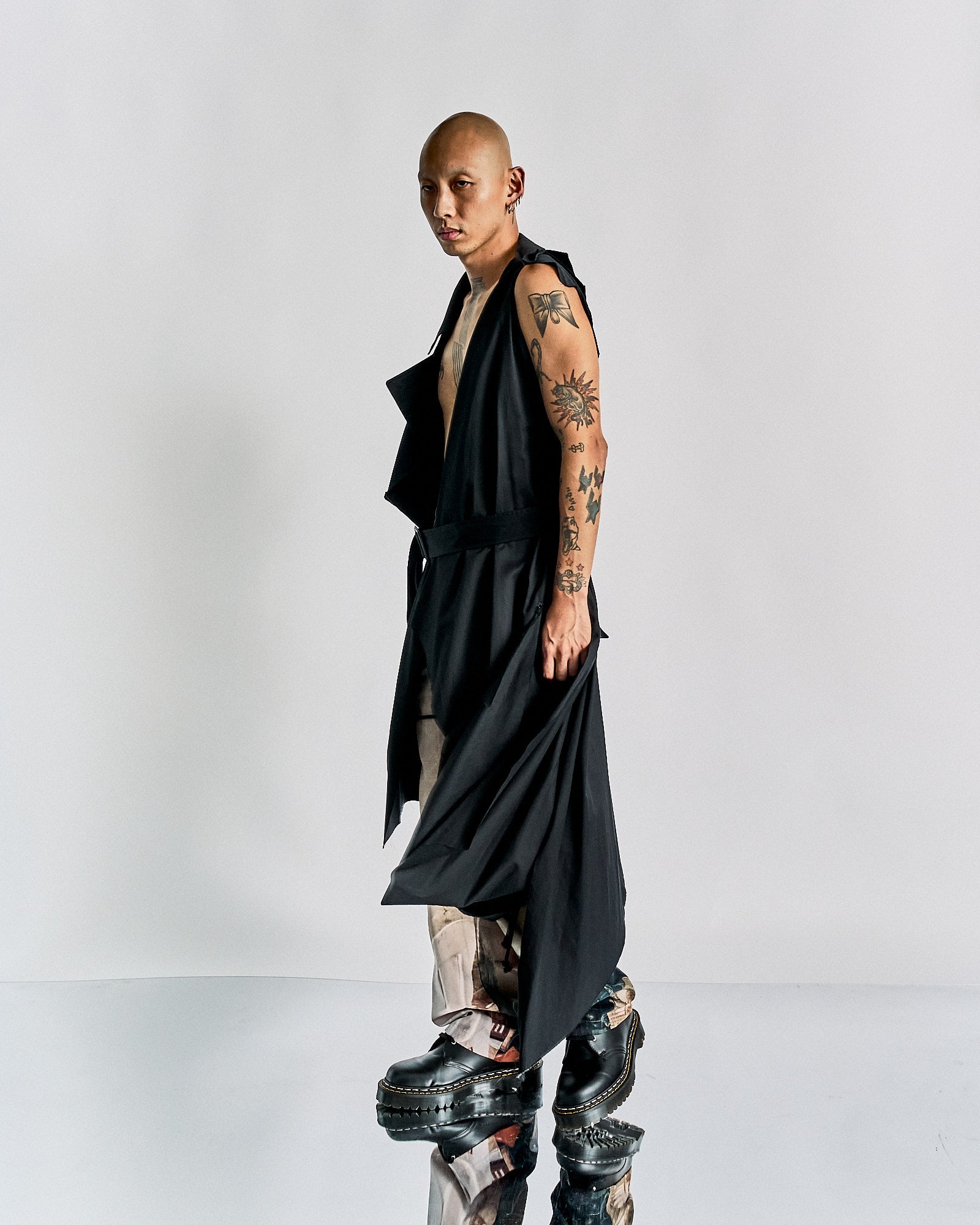 Bald person in a long, sleeveless black Maison Margiela jacket with tattoos, stands on a reflective surface. They wear chunky shoes