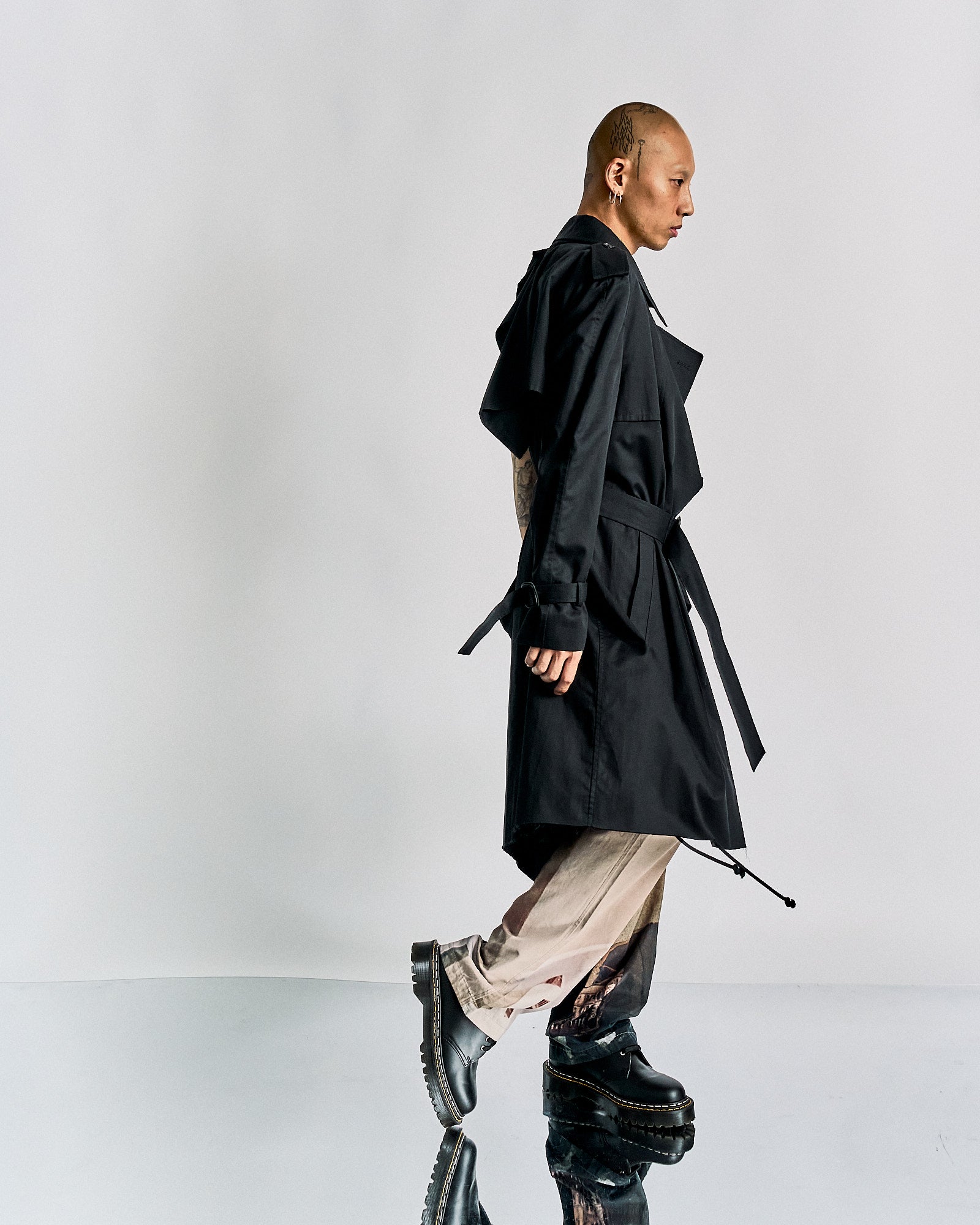A person walks confidently, wearing a long black Maison Margiela trench coat, patterned pants, and black boots against a plain white backdrop, conveying a modern, stylish vibe.