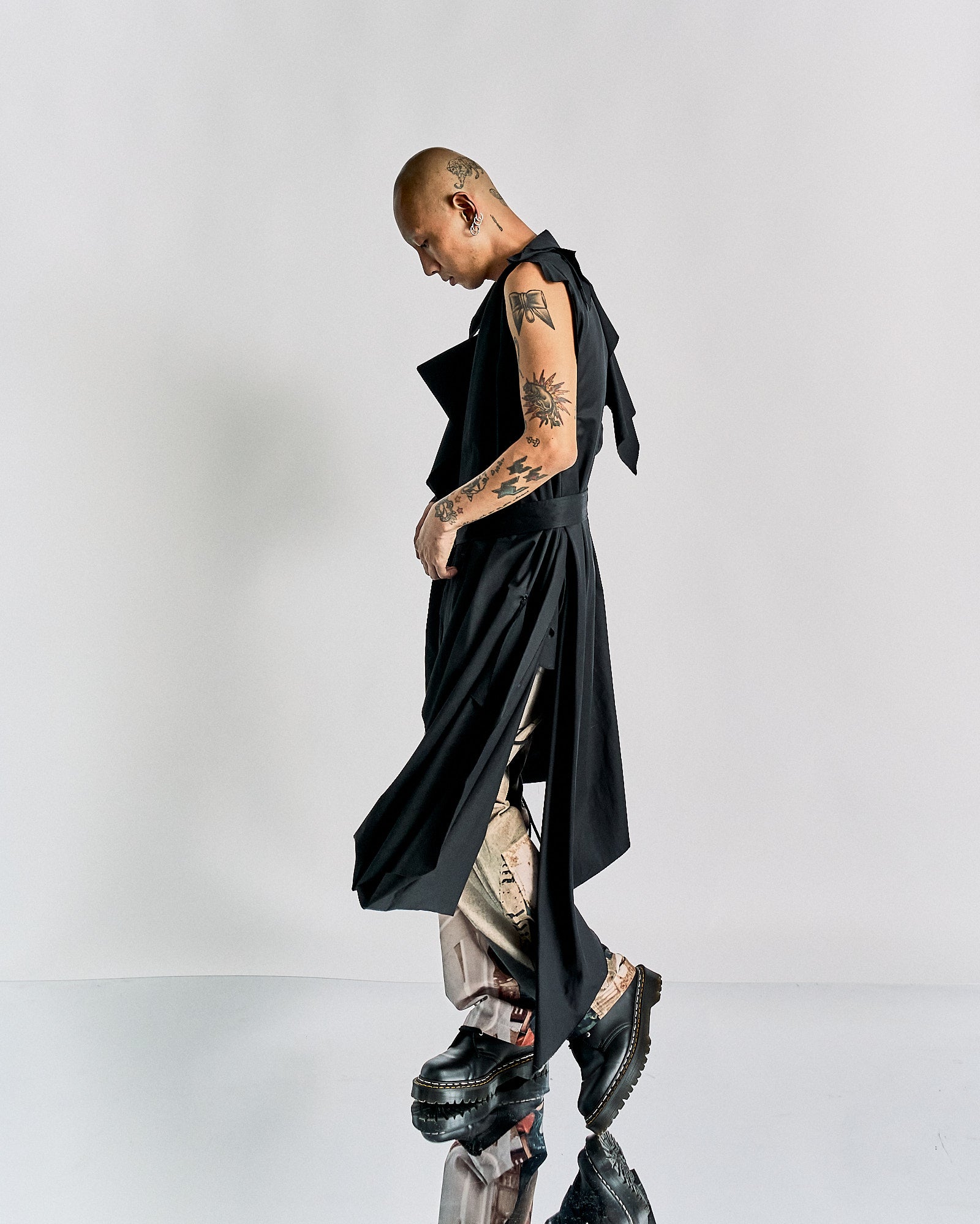 A person with a shaved head and tattoos wears a long, black sleeveless Maison Margiela coat over patterned pants and chunky boots, posing confidently on a reflective floor.