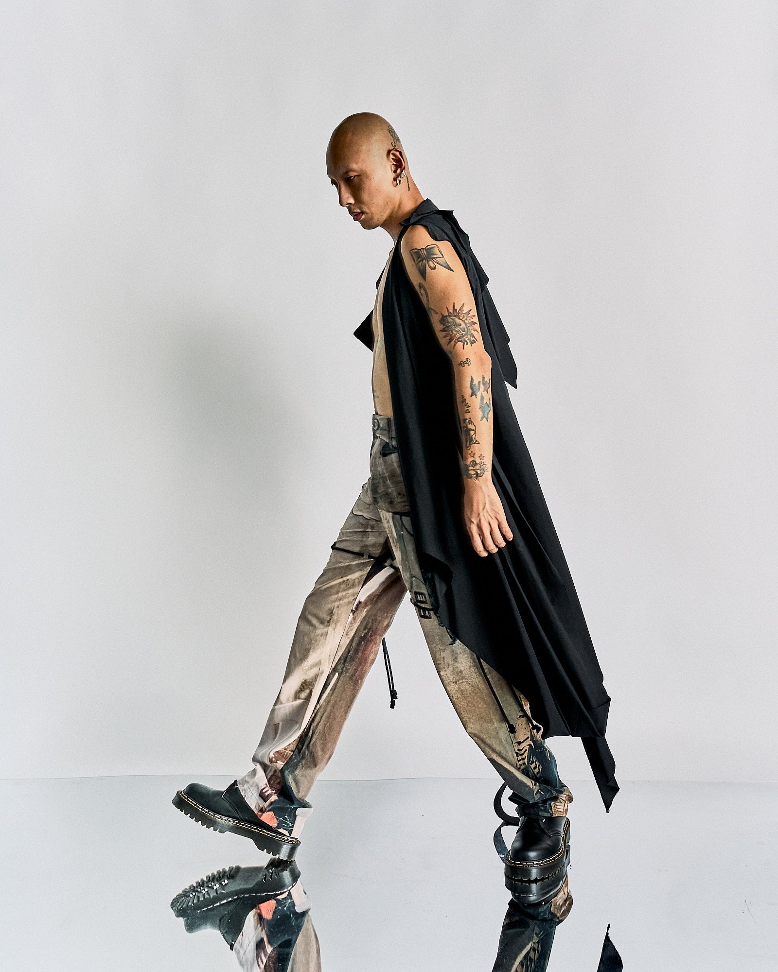 Man walking in profile wearing a sleeveless black Maison Margiela cloak, beige distressed pants, and chunky black shoes. His arms are tattooed, and the background is plain white.