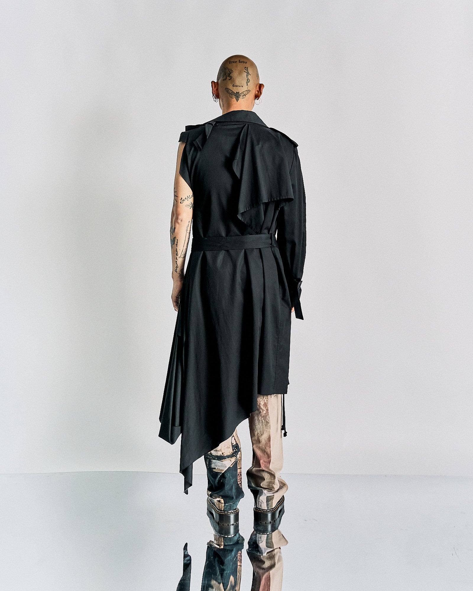 A person stands facing away, wearing an asymmetrical black Maison Margiela outfit and silver boots. Their head and arms display tattoos, conveying a bold, avant-garde style.