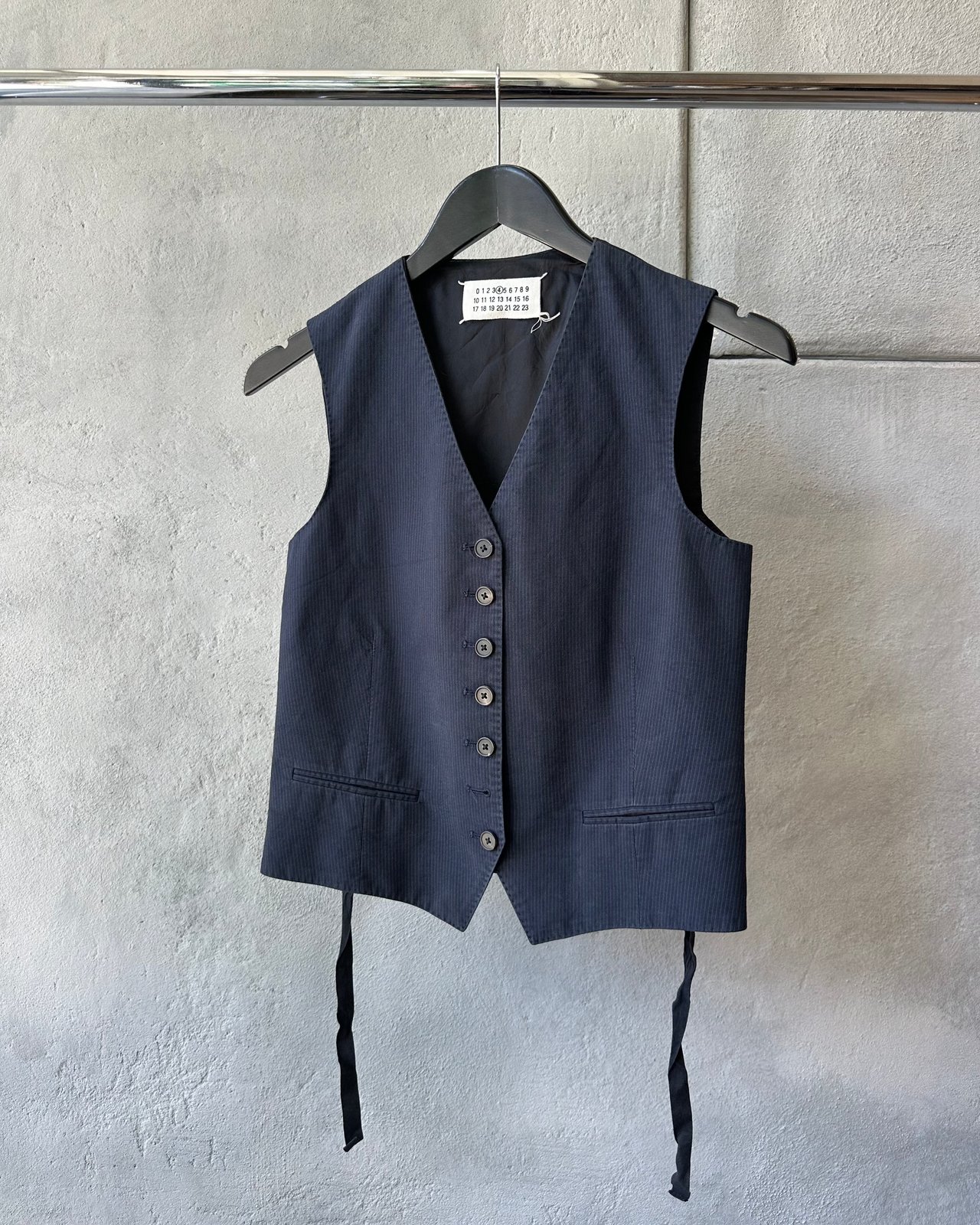 Navy blue vest Maison Margiela with buttons on a hanger against a light gray wall.