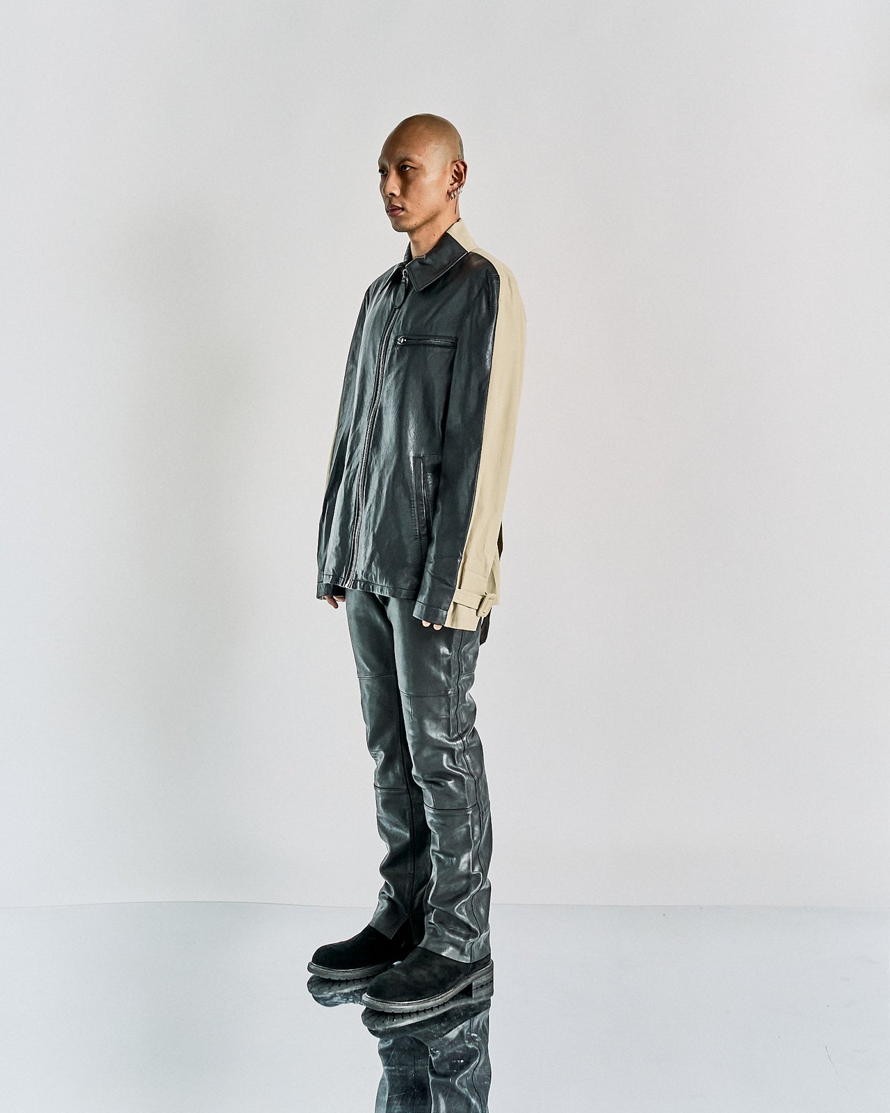 A male model wearing a Maison Margiela leather front zip jacket in black and beige, with a camp-collar and front zip closure, standing in a studio setting.