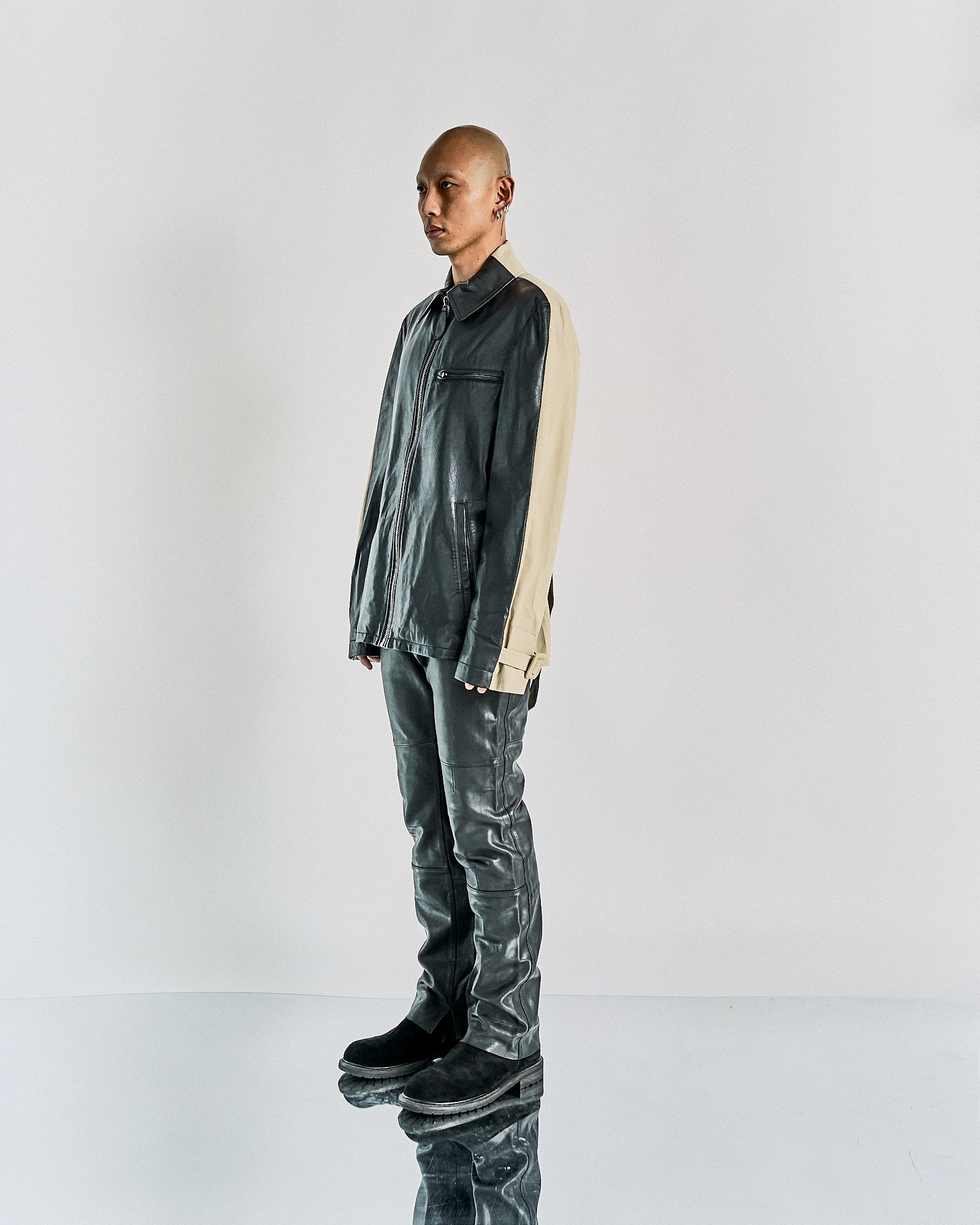 A male model wearing a Maison Margiela leather front zip jacket in black and beige, with a camp-collar and front zip closure, standing in a studio setting.