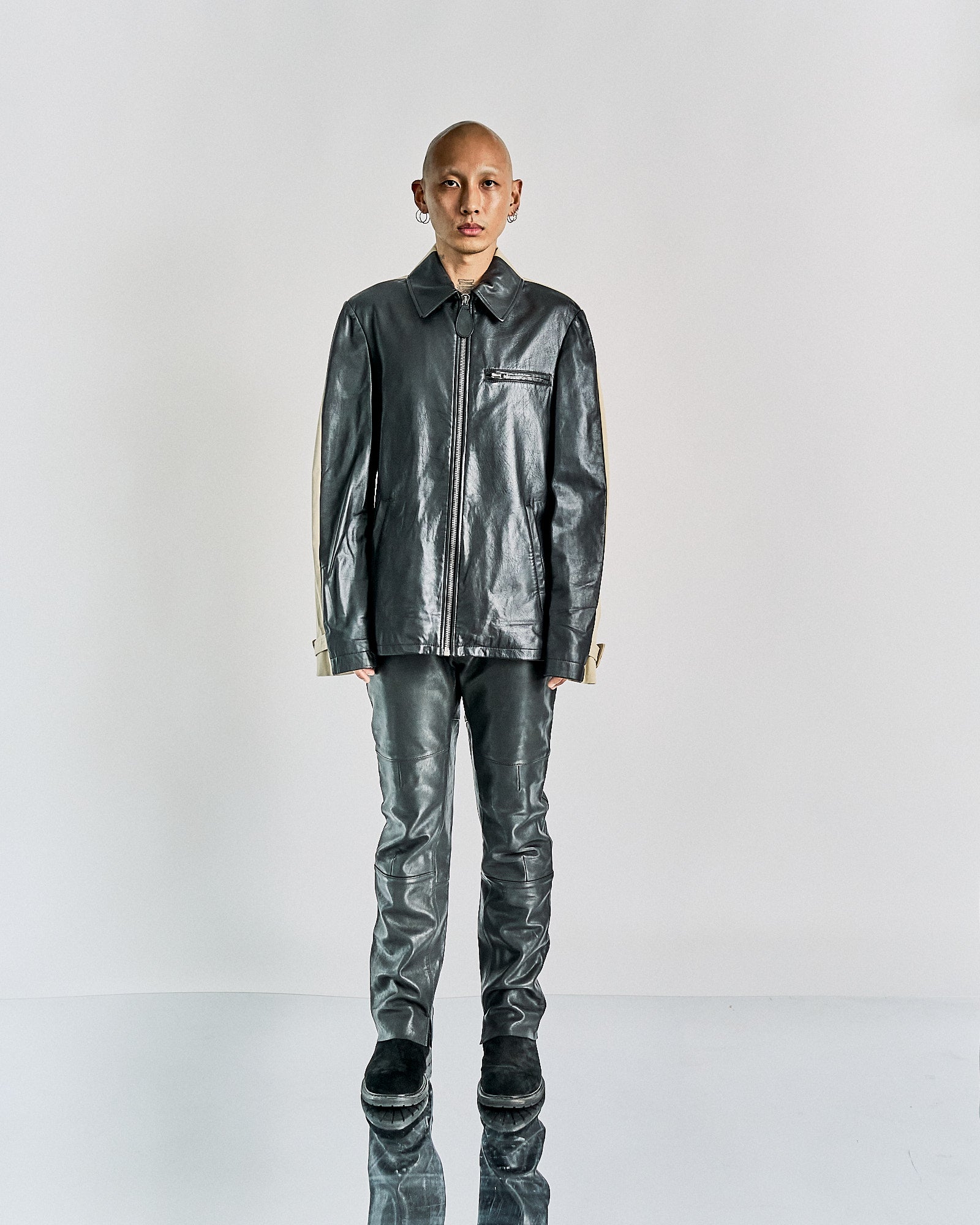 A person stands on a reflective surface wearing a black leather Maison Margiela jacket and pants.