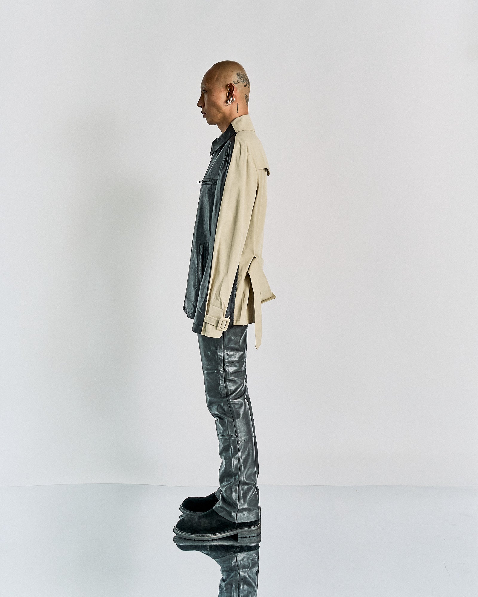 Profile view of a person in a fashion-forward outfit, wearing a layered beige and black Maison Margiela jacket, black leather pants, and black shoes. The background is plain white.
