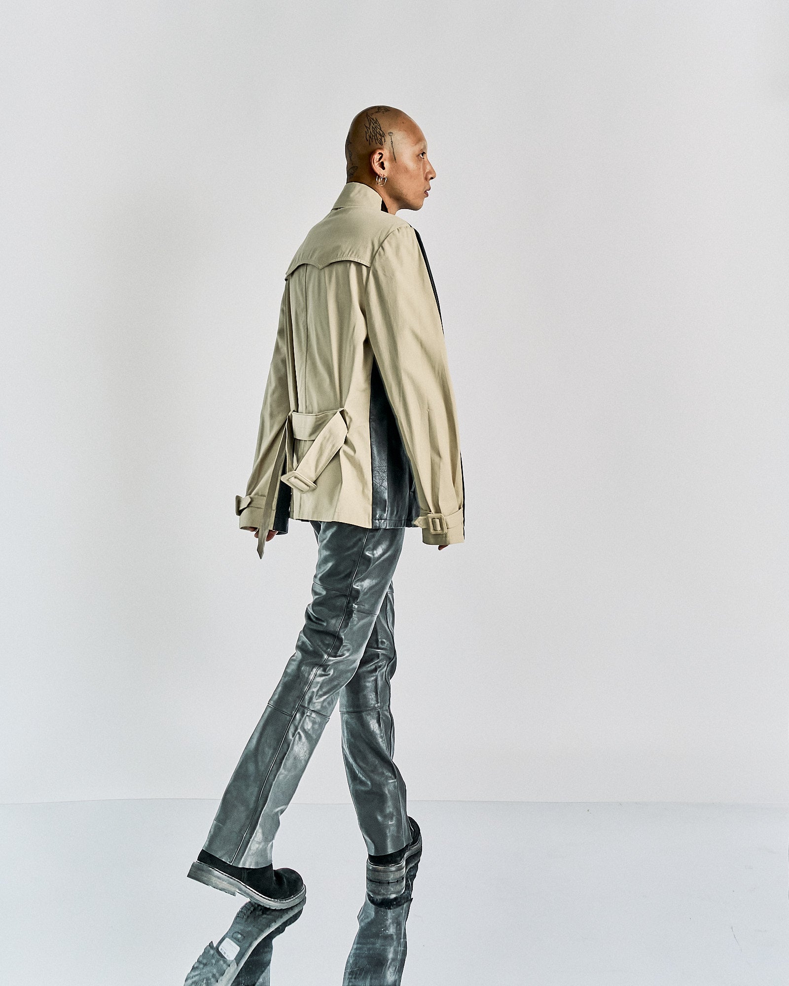 Person with a shaved, tattooed head seen from behind, wearing a beige Maison Margiela jacket with a belt and shiny grey pants, standing against a plain background.