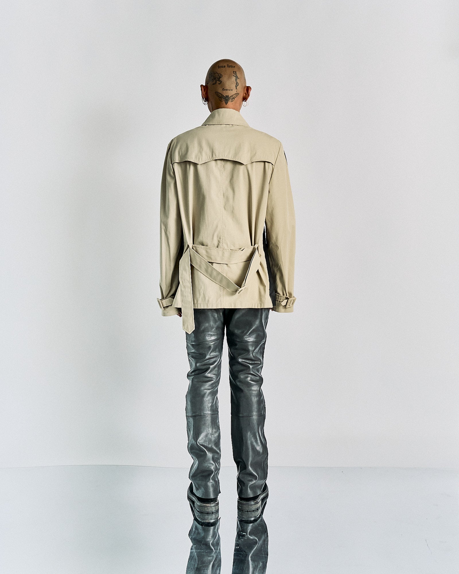 Person with a shaved, tattooed head seen from behind, wearing a beige Maison Margiela jacket with a belt and shiny grey pants, standing against a plain background.
