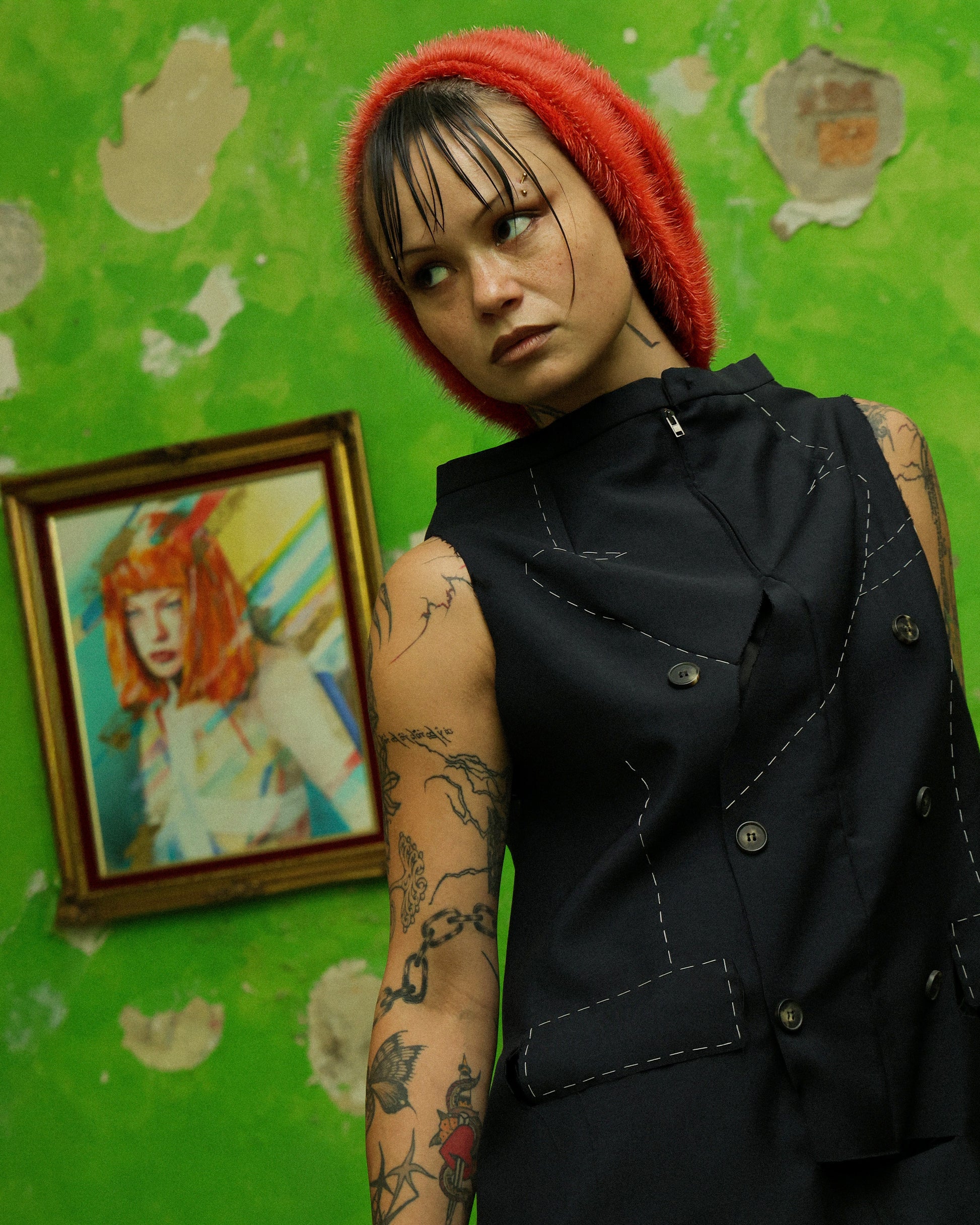 A model wearing a black Maison Margiela top hybrid with a wool mohair skirt, featuring a zip collar, double-breasted front, and cut-out details, accessorized with a red headpiece, standing in front of a green textured wall with a portrait painting.