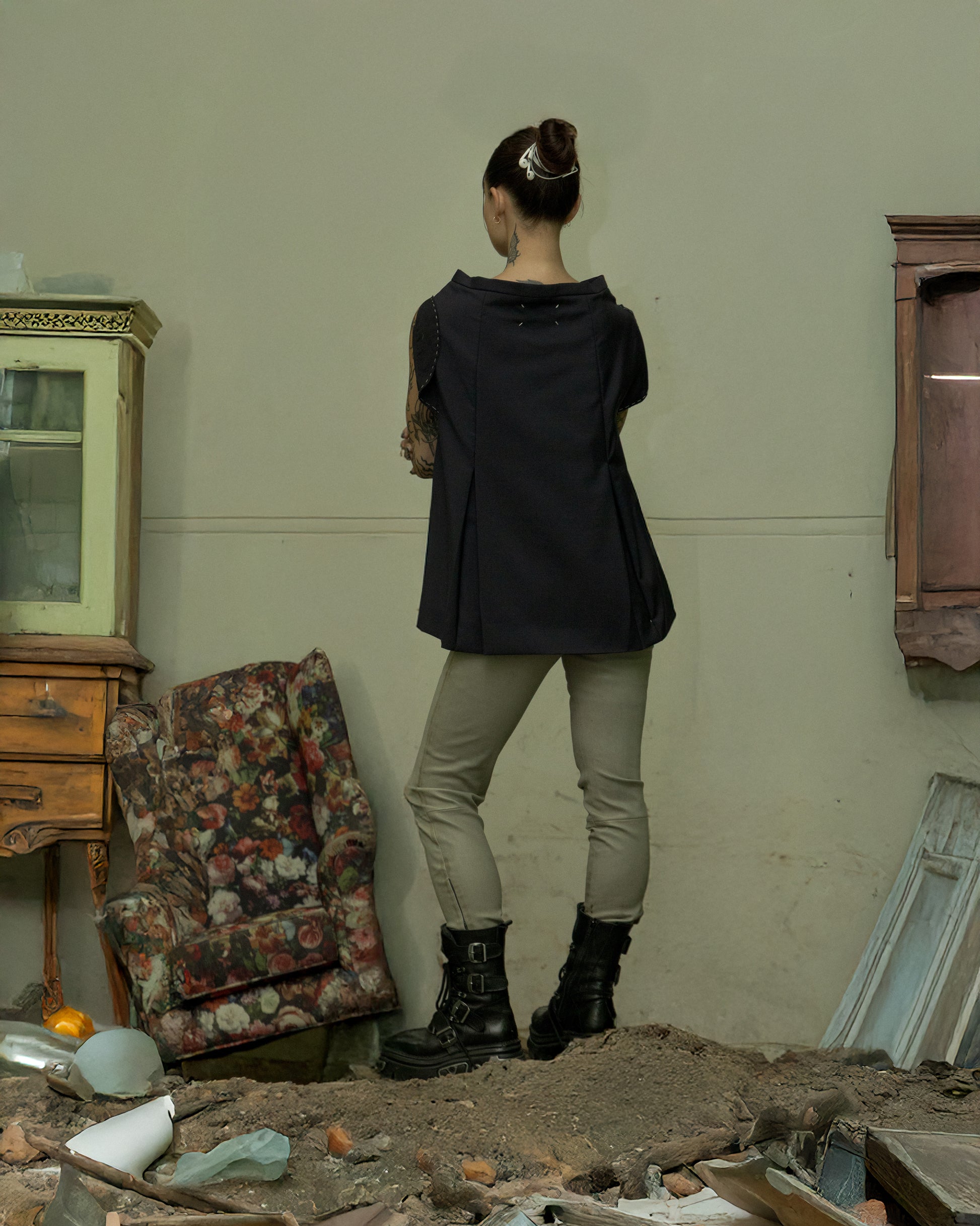 Person wearing a black Maison Margiela top and beige pants standing in a room with debris and old furniture.