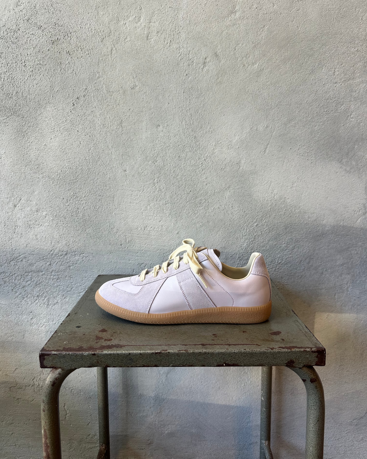 Purple Maison Margiela sneaker with yellow laces on a metal stool against a textured wall.