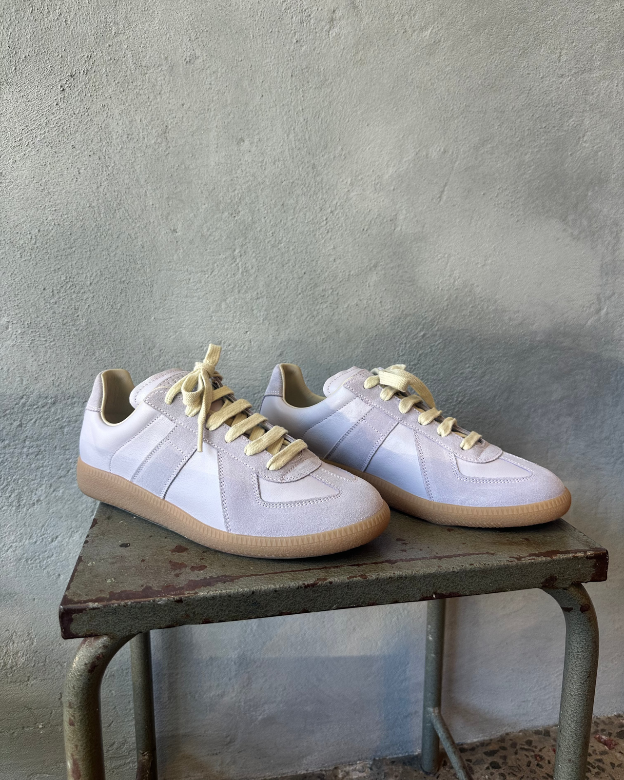 Purple Maison Margiela sneaker with yellow laces on a metal stool against a textured wall.