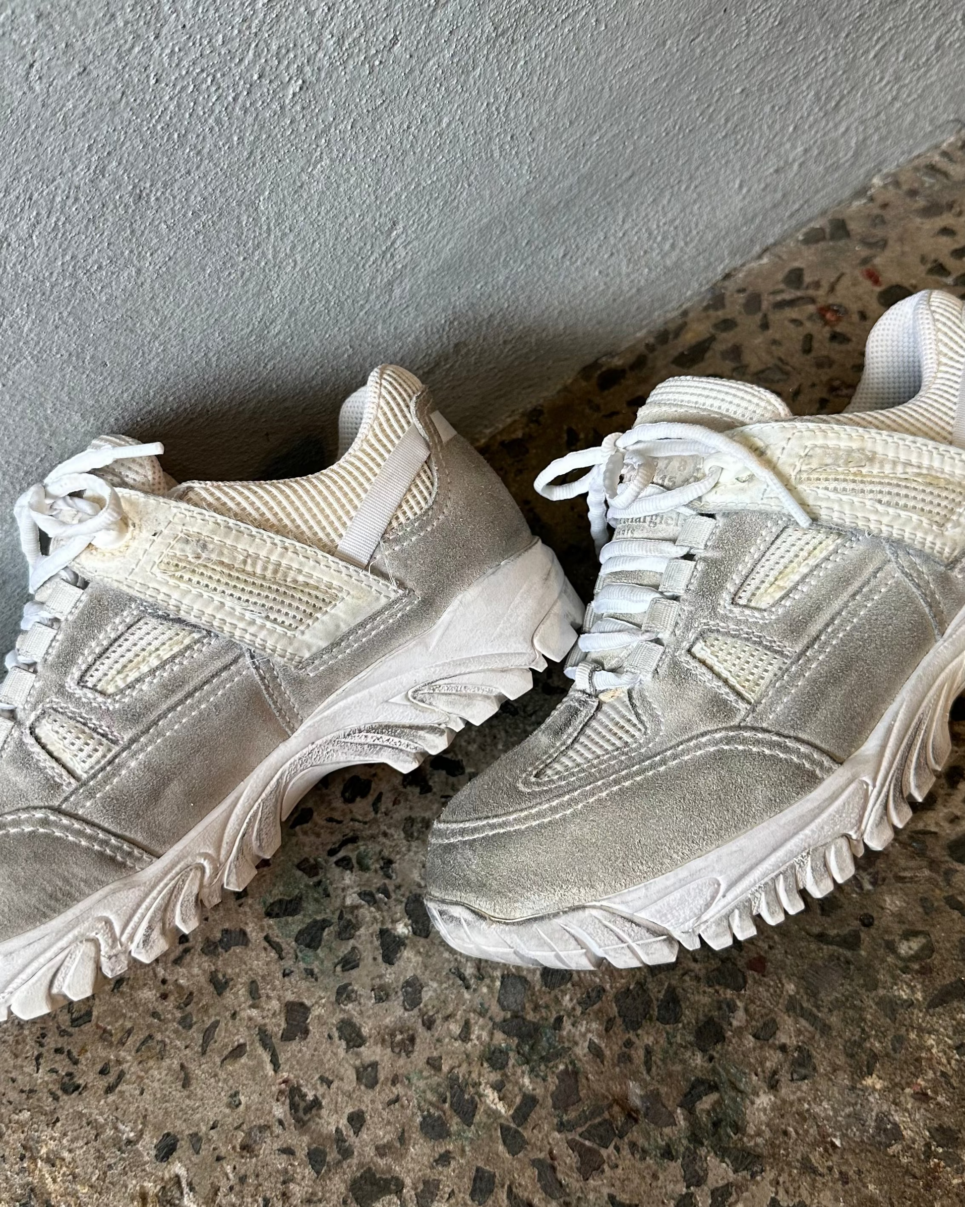 Pair of light gray Maison Margiela sneakers on a speckled surface with a neutral wall background