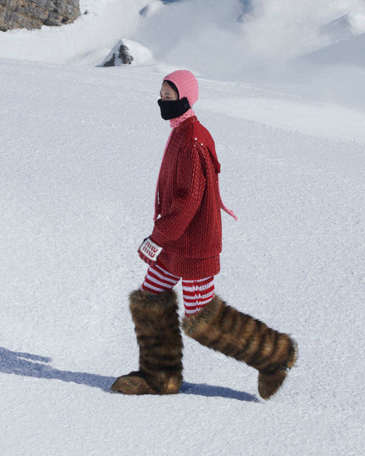 Person in red Miu Miu coat and striped pants walking on a snowy landscape with mountains in the background