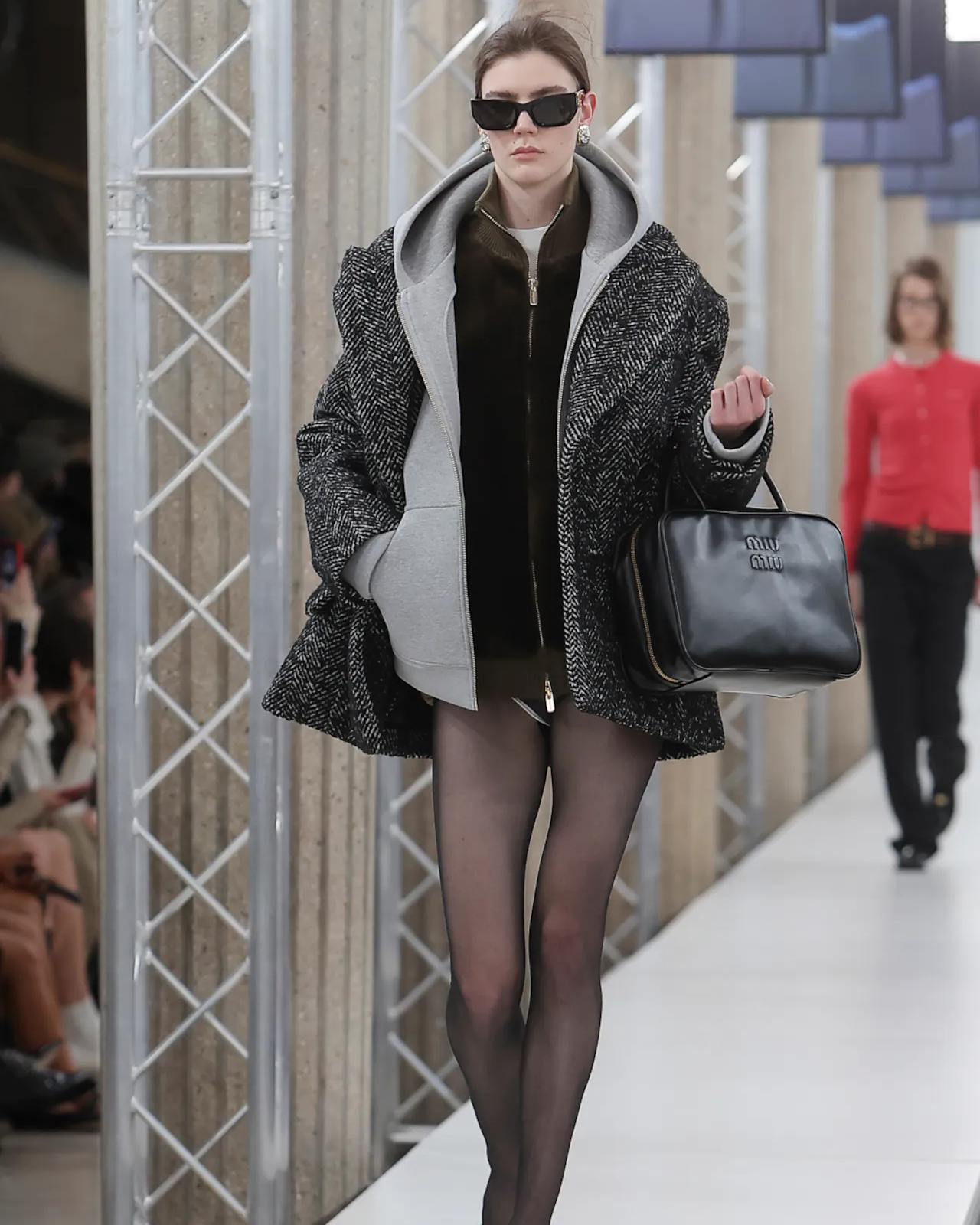 Model walking on a Miu Miu catwalk wearing a stylish outfit with a coat and sunglasses.
