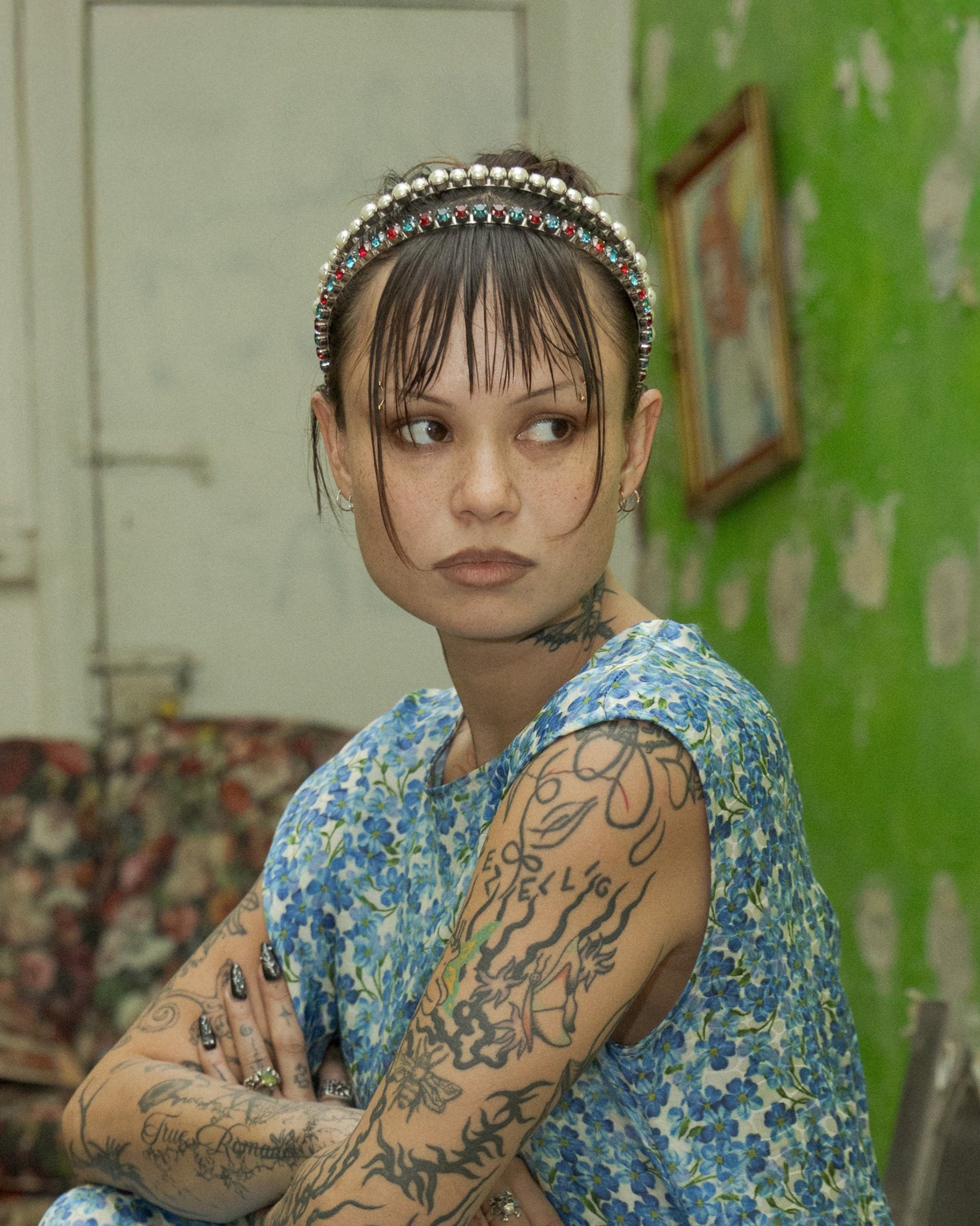 Person with tattoos wearing a Miu Miu double headband sitting in a room with green wallpaper and books on the floor.