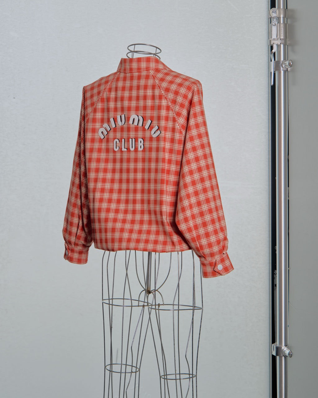 Red checkered Miu Miu bomber jacket with 'Miu Miu Club' text on a mannequin against a grey background