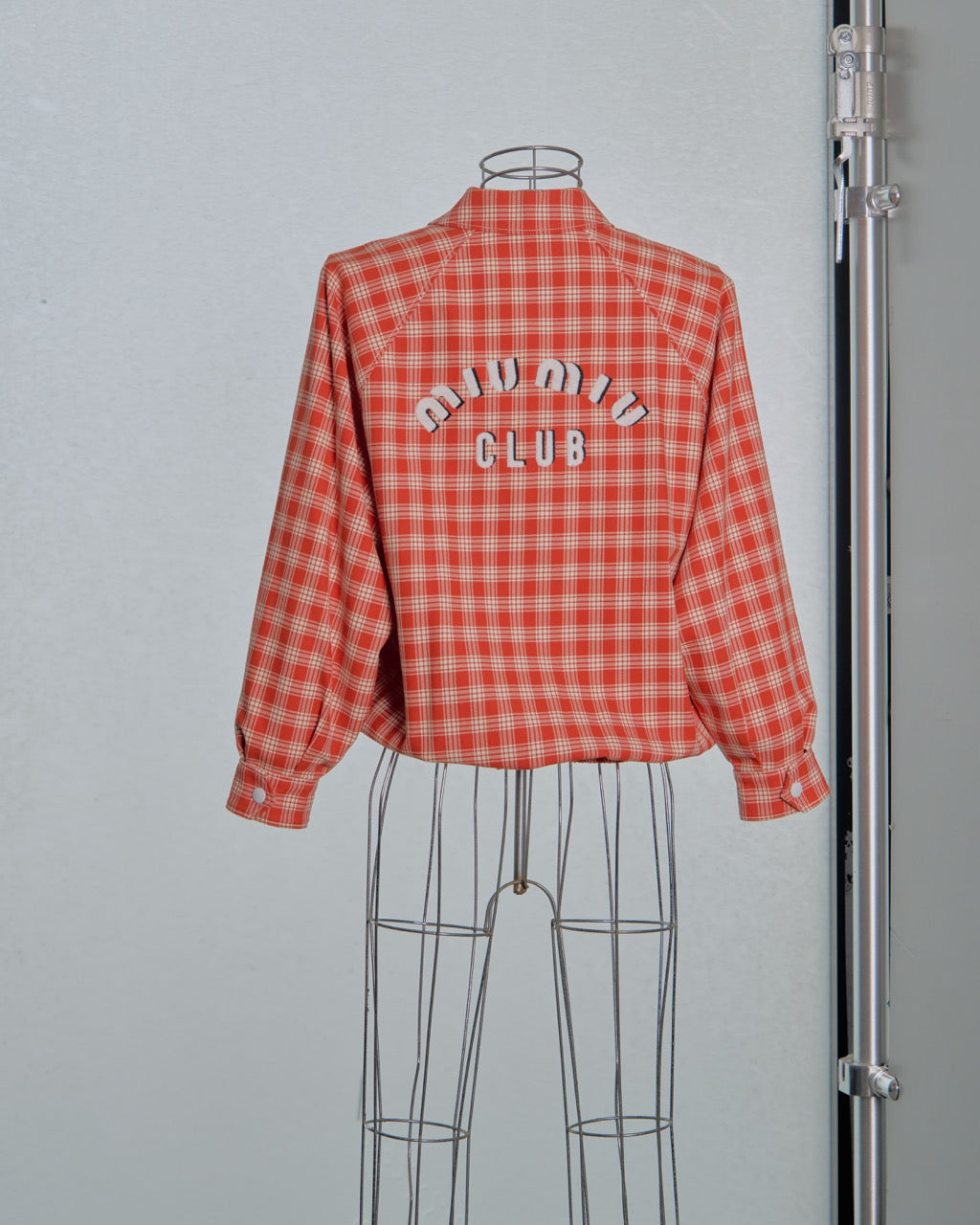 Red and white checkered Miu Miu bomber jacket with 'Miu Miu Club' text on a mannequin against a gray background