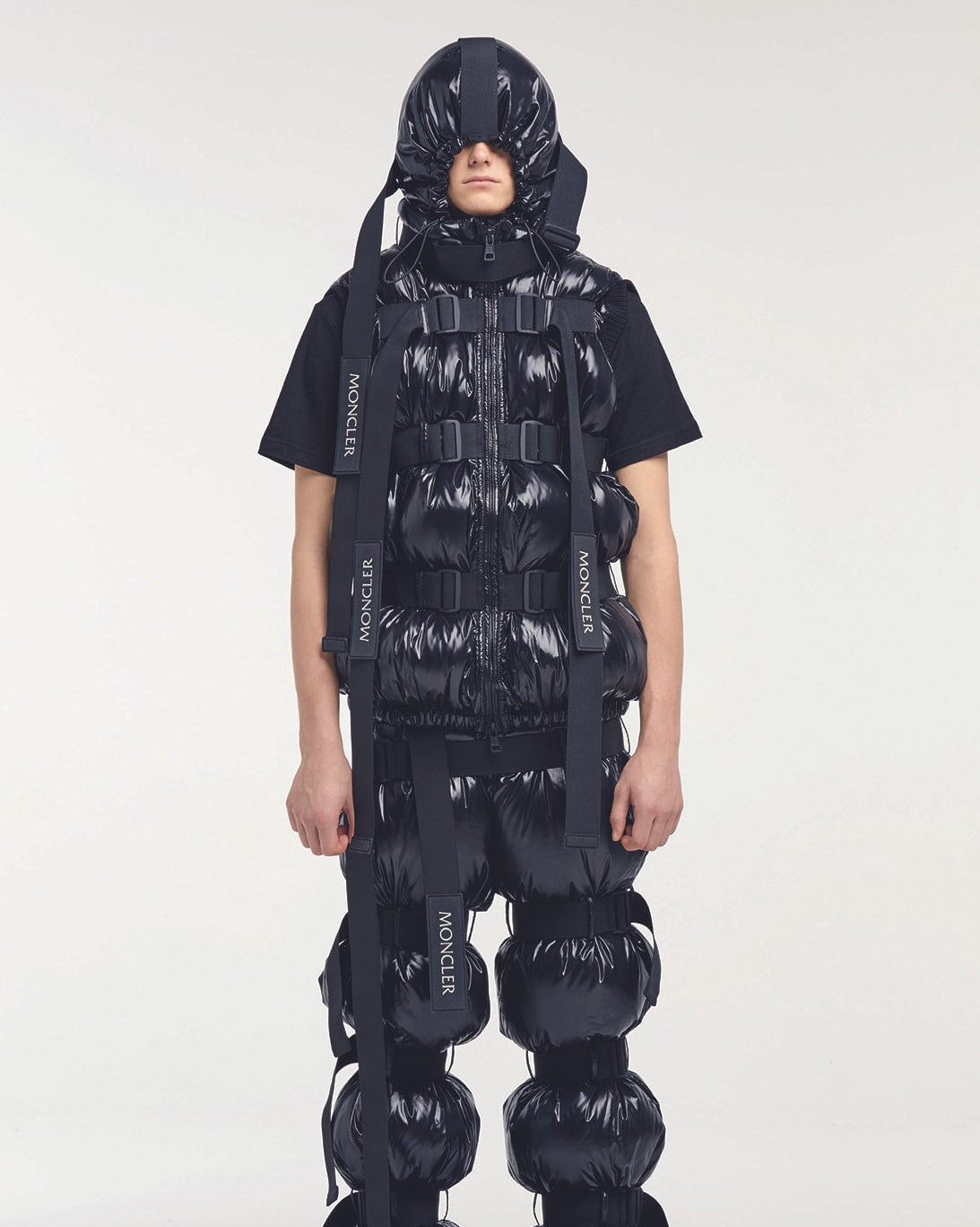 Person in futuristic, padded Moncler x Craig Green black outfit with a hood and layered puffy sections. Stands against a neutral background.