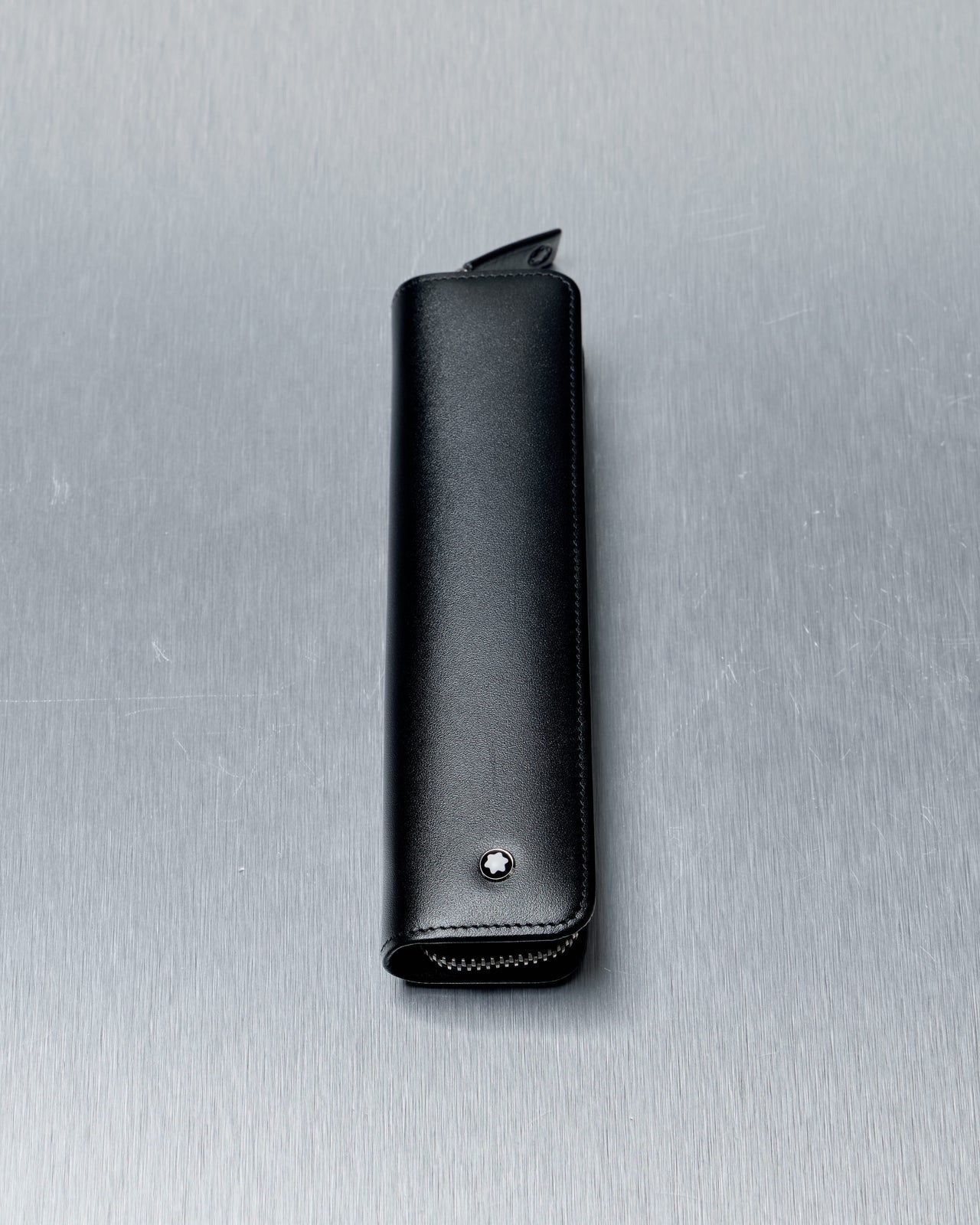 Black leather Mont Blanc pen case with a zipper and a small star emblem, set against a textured, metallic background.