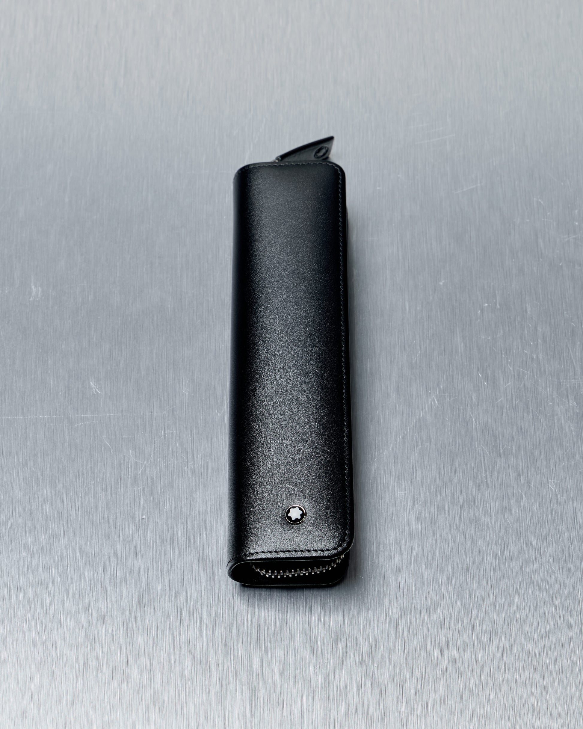 Black leather Mont Blanc pen case with a zipper and a small star emblem, set against a textured, metallic background.
