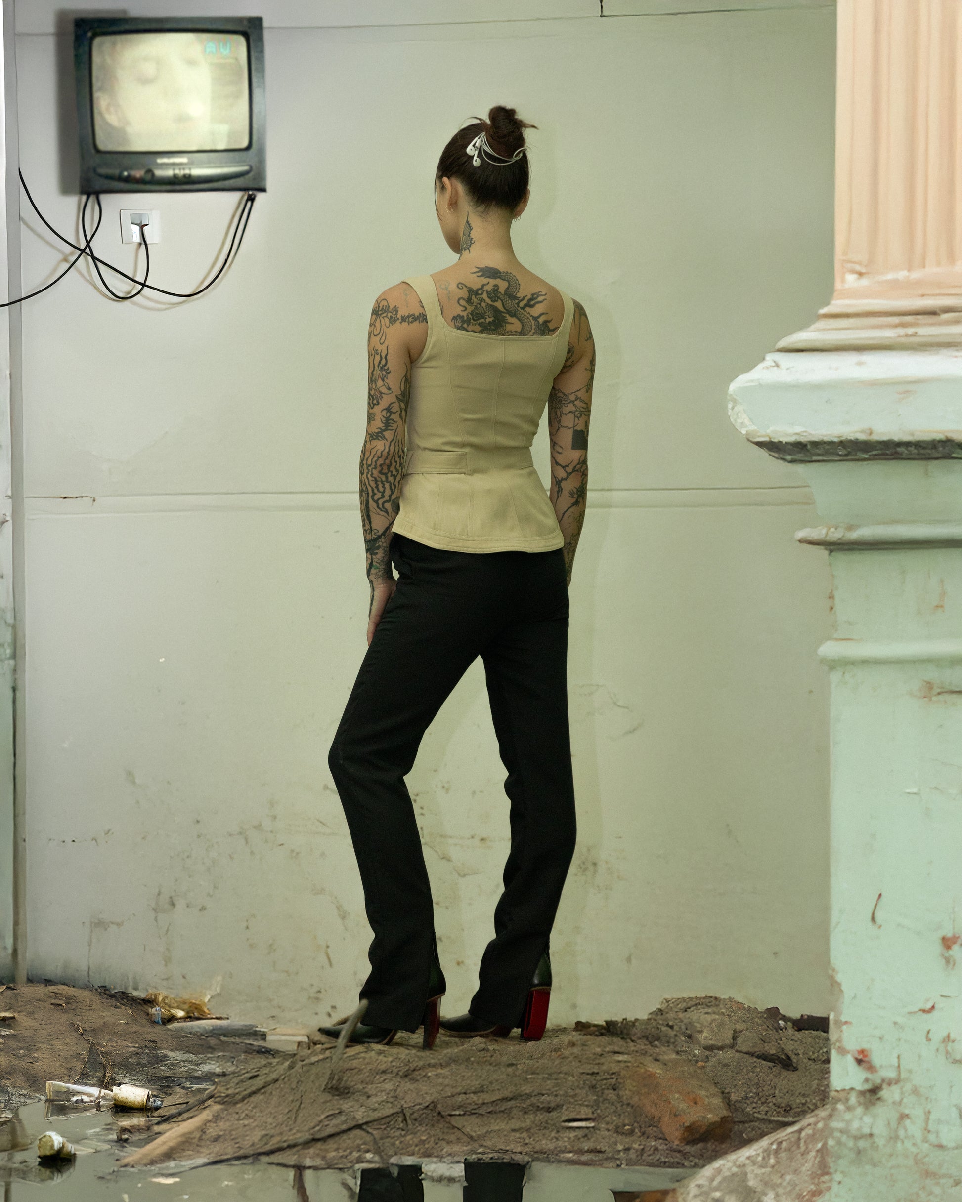 Person with tattoos standing in a room with a television on the wall wearing a beige Mugler top.