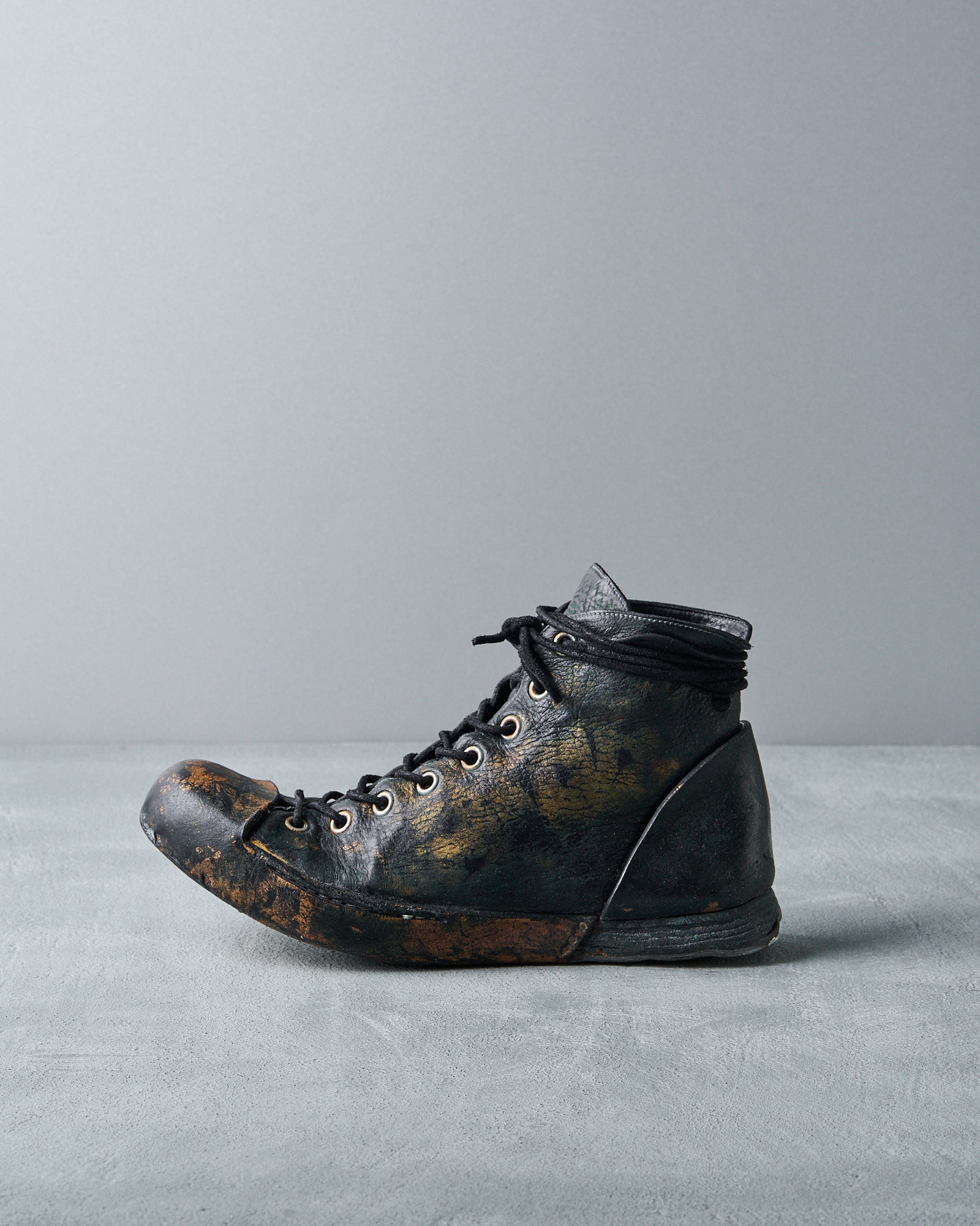 A Nihomano black boot with visible scuffs and creases. It features laces threaded through metal eyelets and sits on a plain grey background. 