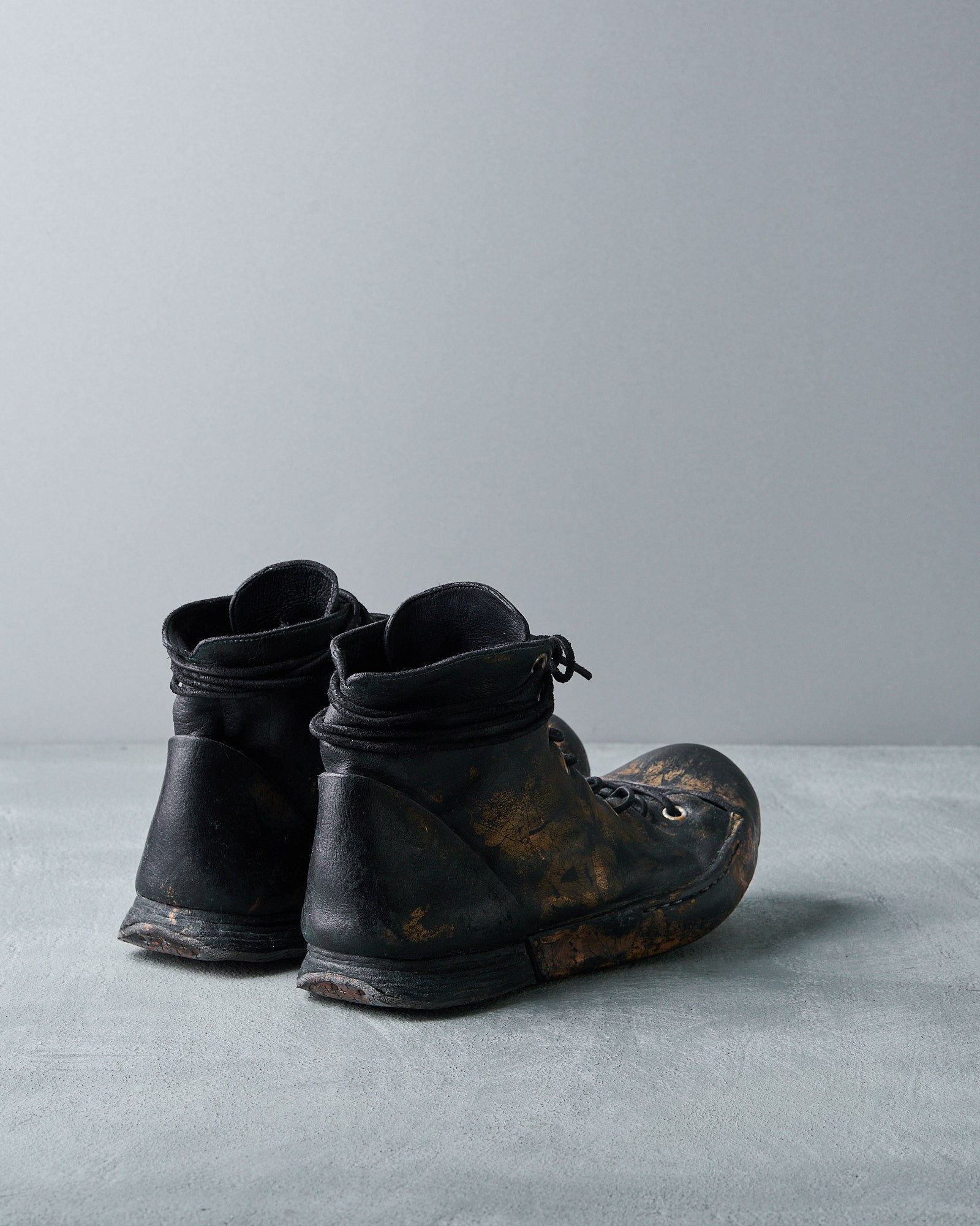 Black leather Nihomano boots with textured, dirt-stained surfaces, resting on a matte grey surface 