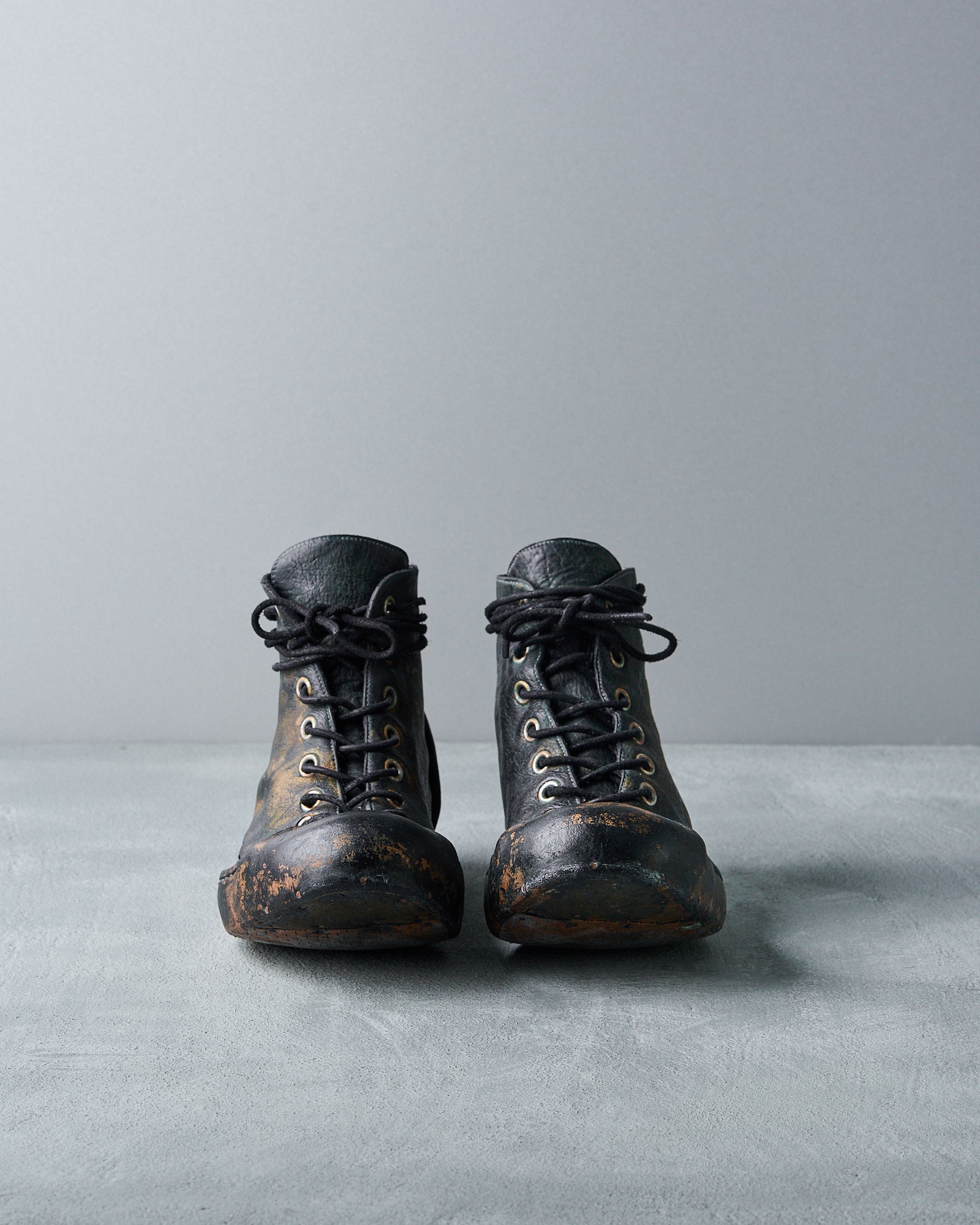 Black Nihomano boots with laces and scuffed toes are standing on a light grey surface against a plain grey background