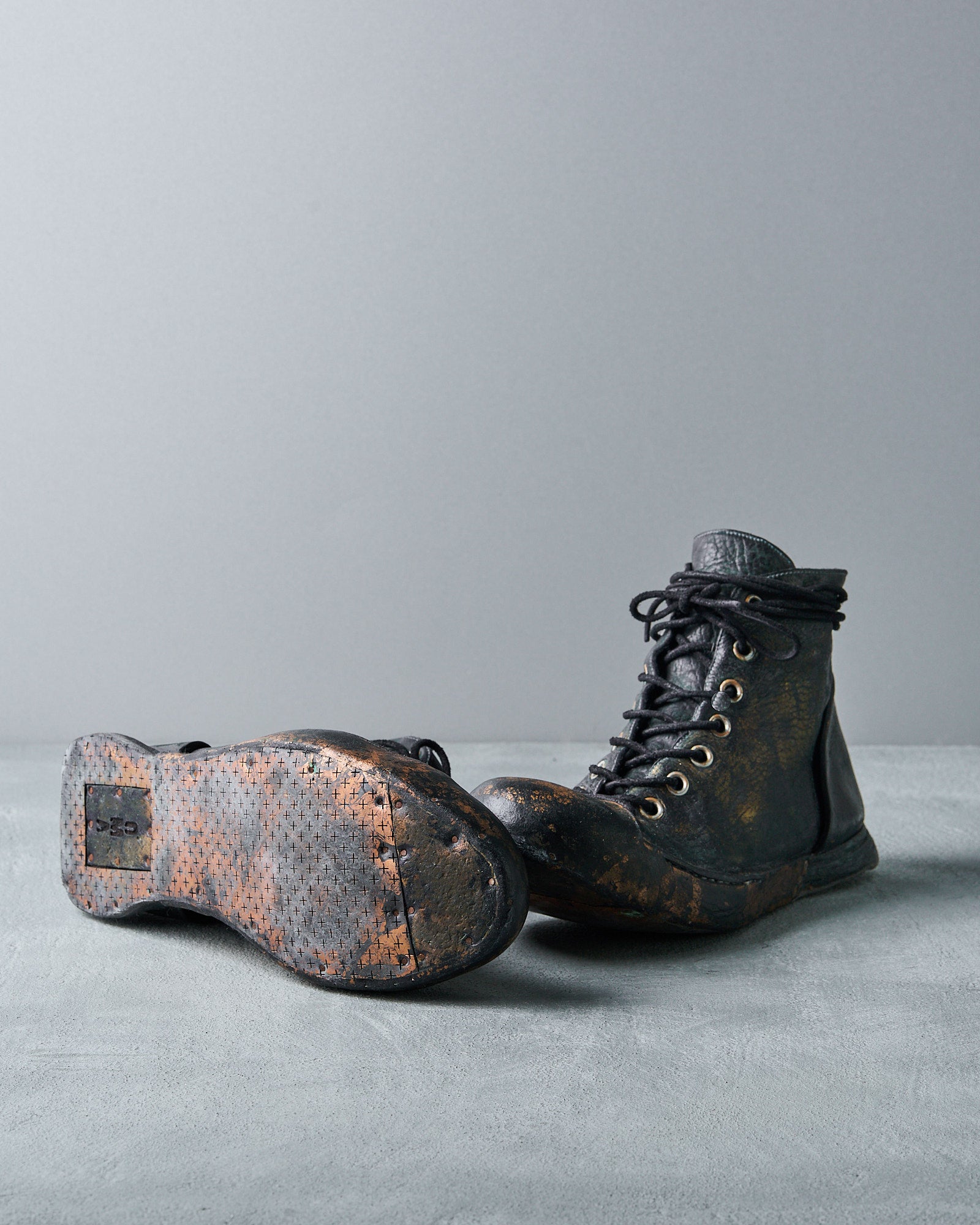Black Nihomano lace-up boots with scuffed soles placed on a textured surface. One boot is upright, the other lies on its side