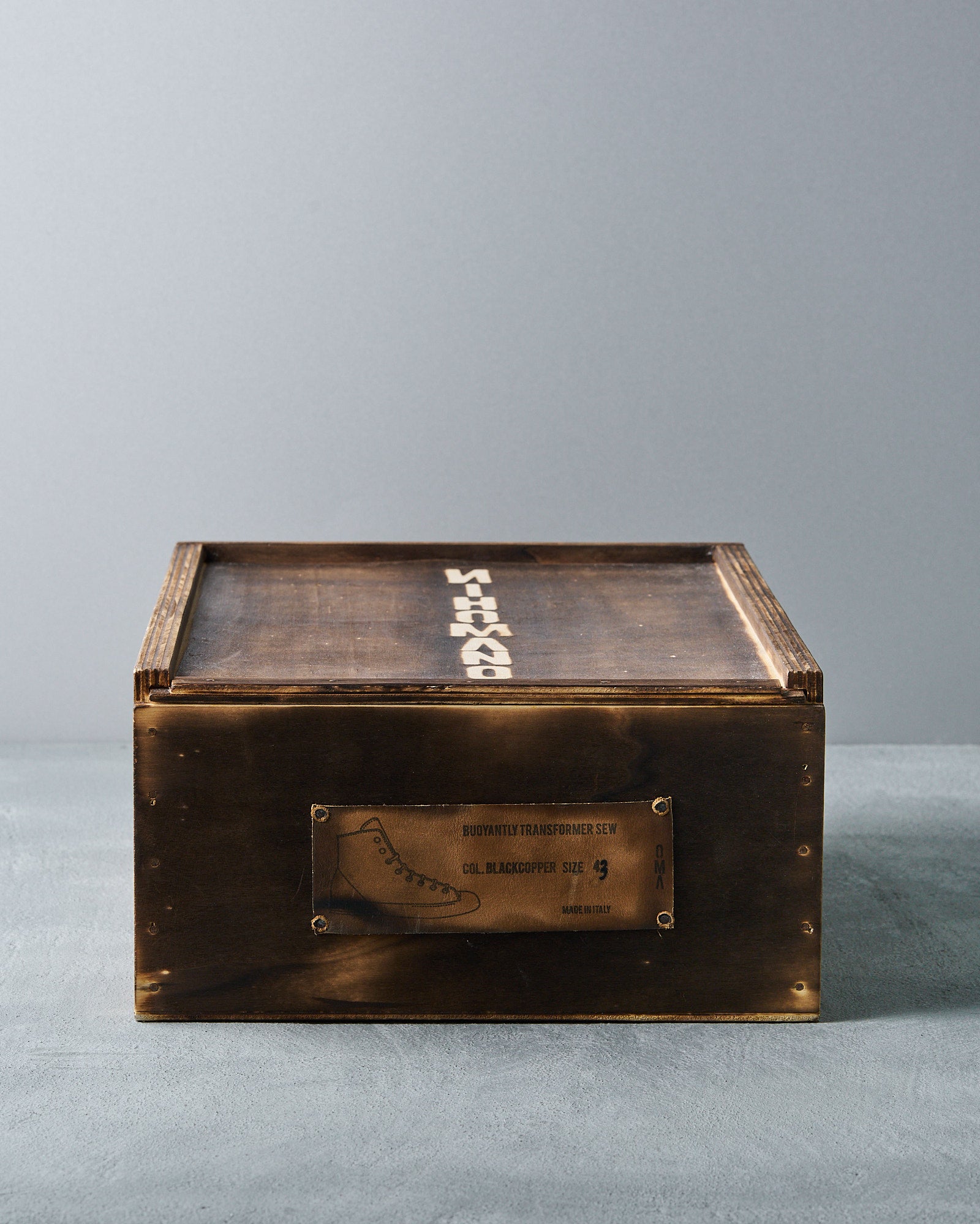 Wooden Nihomano box with dark, weathered finish, featuring bold, white text on top. A leather patch with shoe details is attached