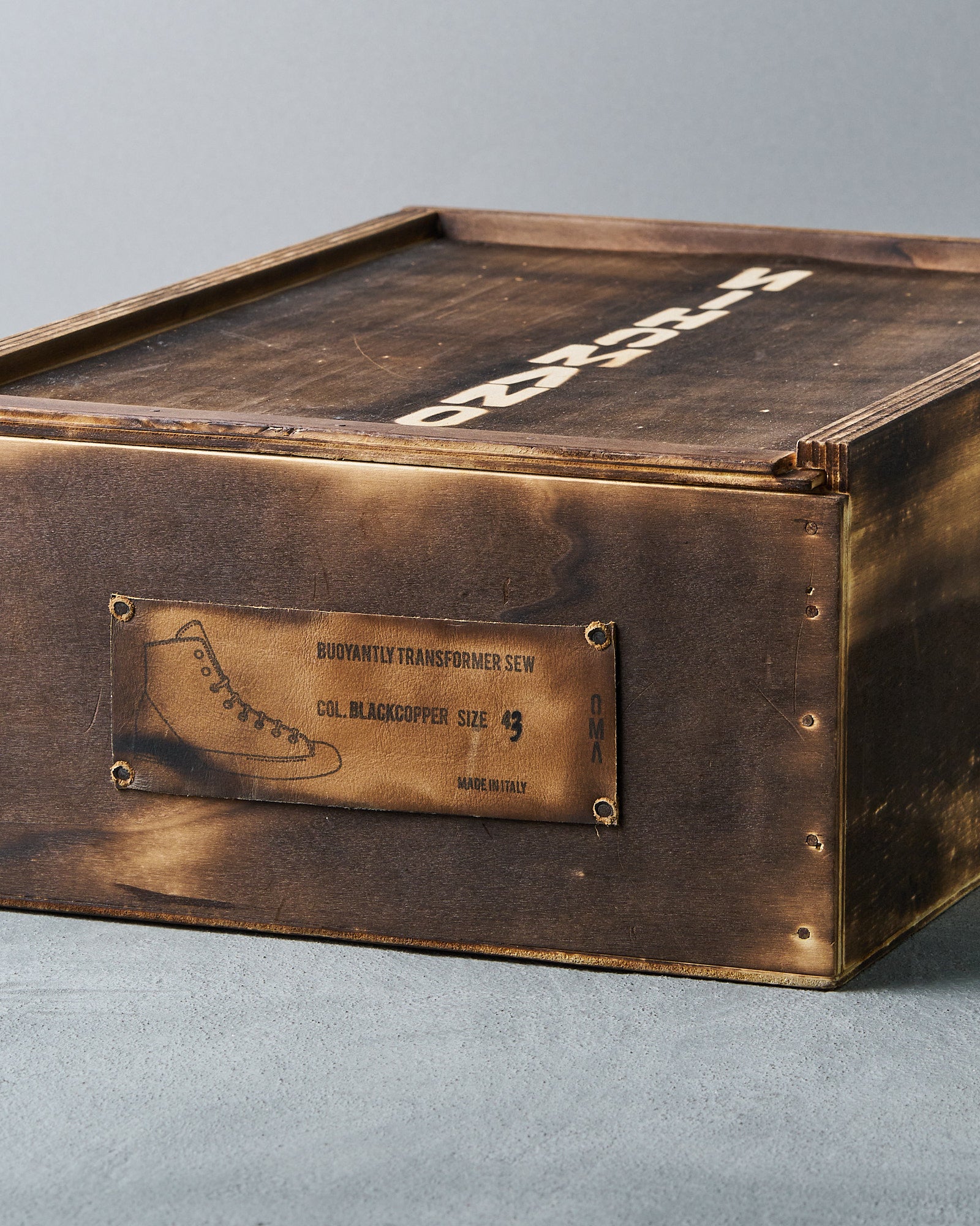 A rustic brown wooden Nihomano shoe box with a stencil-like label on the side, displaying a boot design and text: "Buoyantly Transformer Sew, Blackcopper, Size 43, Made in Italy.”