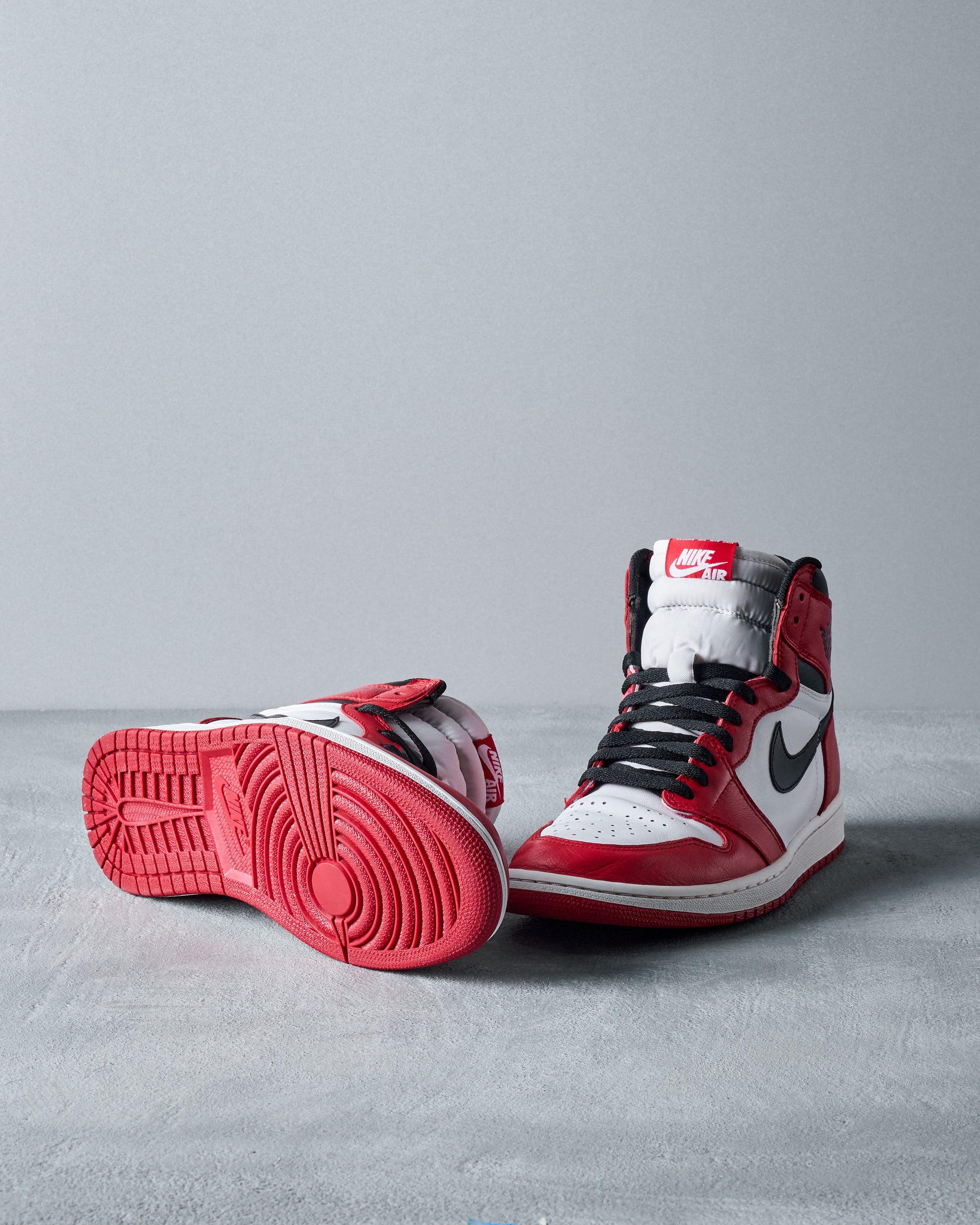 Red, white, and black Nike Air Jordan high-top sneakers are displayed on a gray surface. One shoe is upright, showing the Nike logo, while the other lies on its side, revealing the red sole.