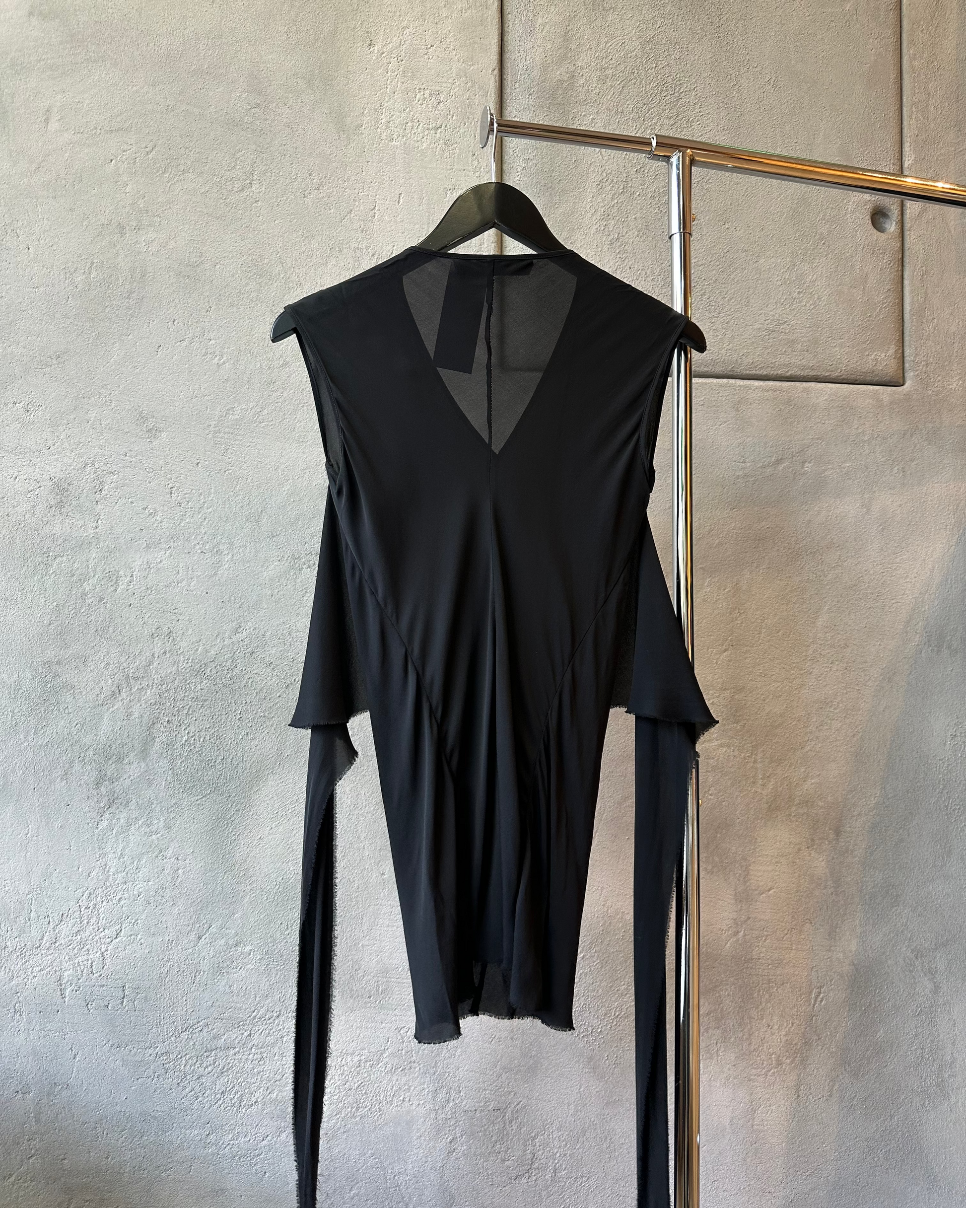 Black sleeveless P.R.Patterson blouse with sheer panels on a hanger against a textured wall.