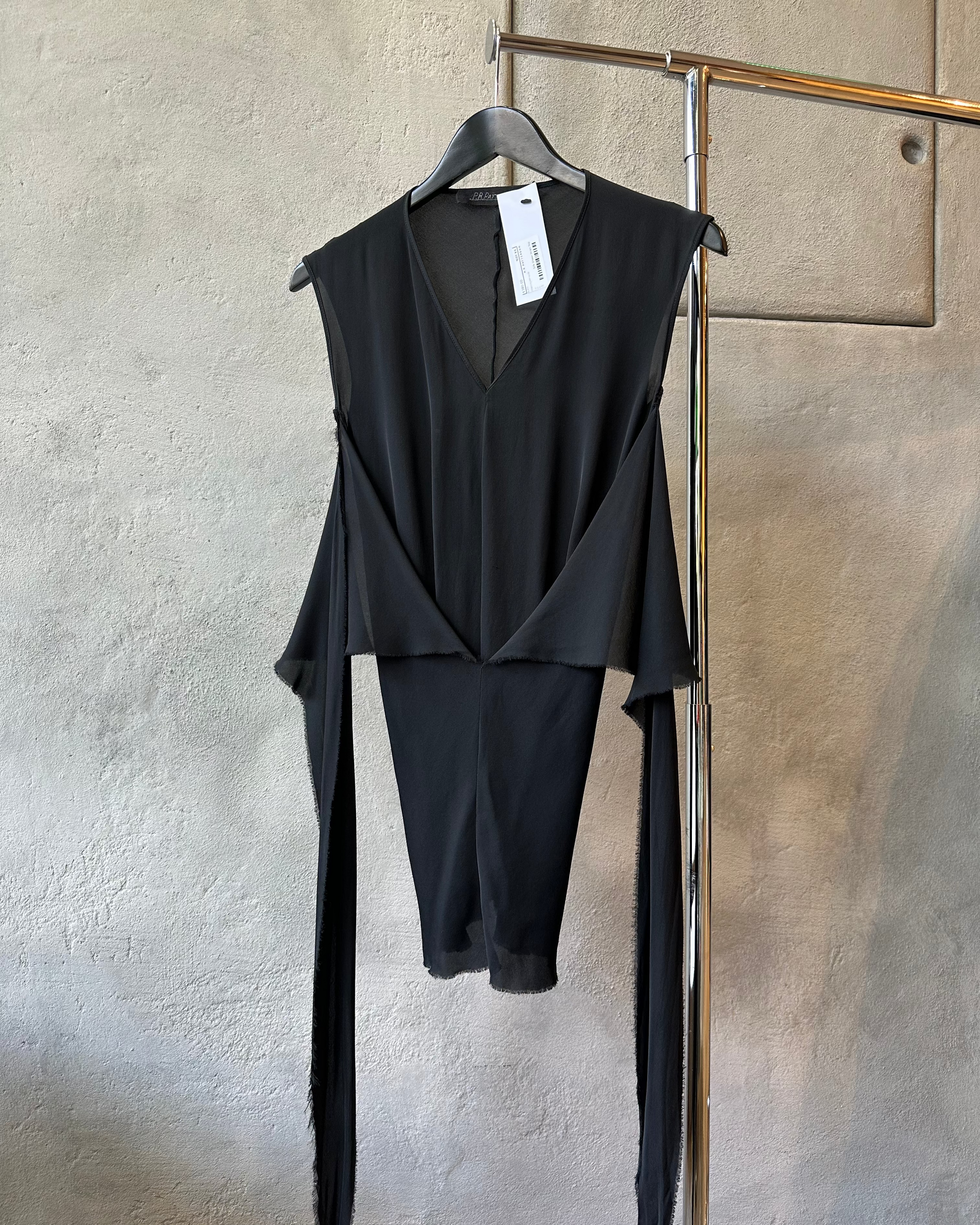 Black sleeveless P.R.Patterson top on a hanger against a textured wall.
