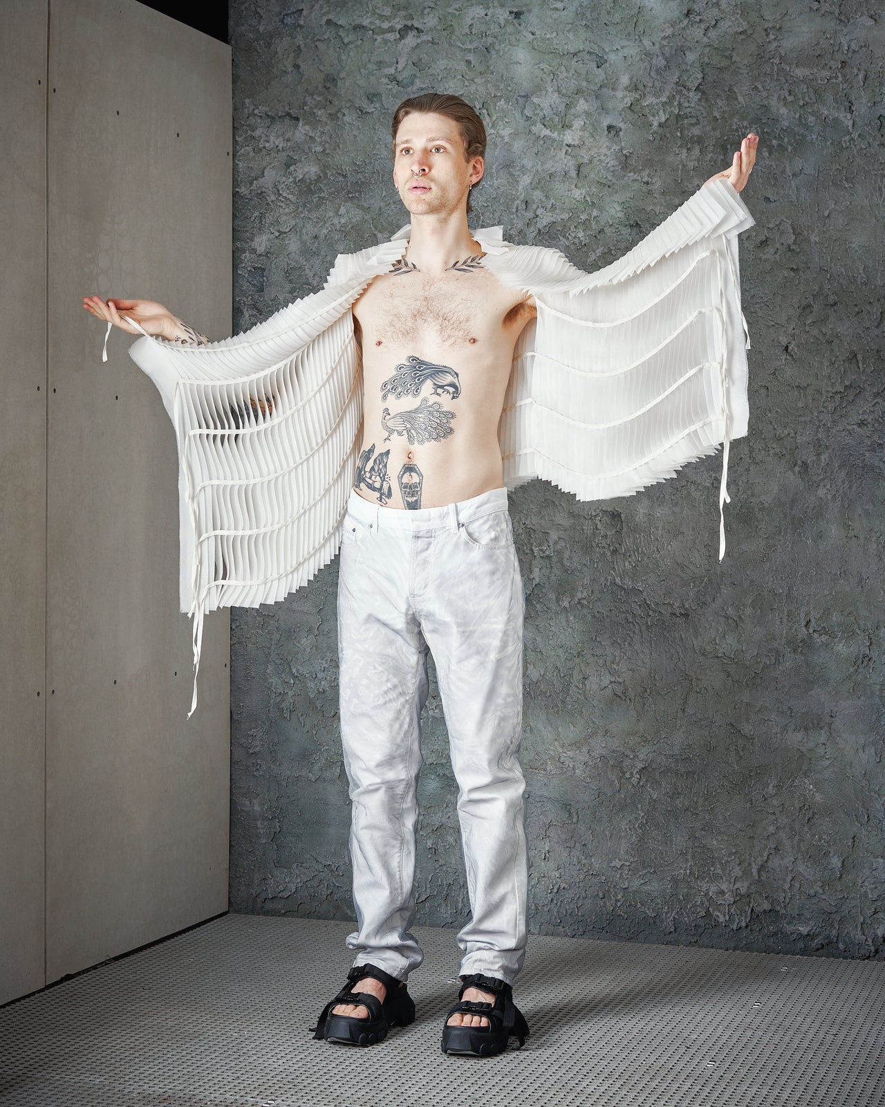 A person standing with their arms raised and spread out, wearing a white, layered silk Peachoo + Krejberg  vest with a geometric pattern and a black tattoo on the chest.