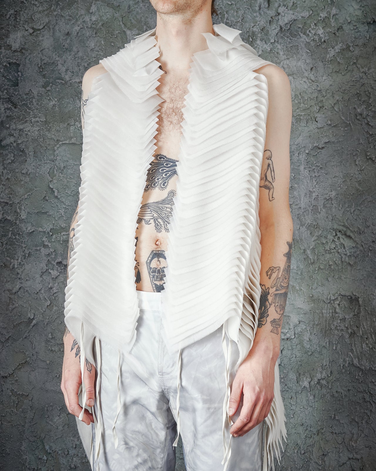 A person standing with their arms raised and spread out, wearing a white, layered silk Peachoo + Krejberg  vest with a geometric pattern and a black tattoo on the chest.