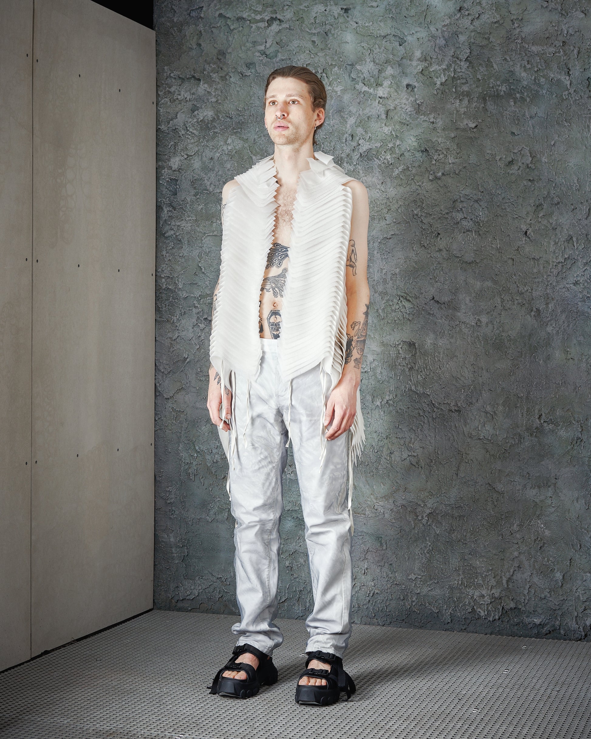 A person stands against a textured gray wall, wearing a white, fringed Peachoo + Krejberg vest and shiny silver pants. Tattoos are visible, and the tone is edgy and stylish.
