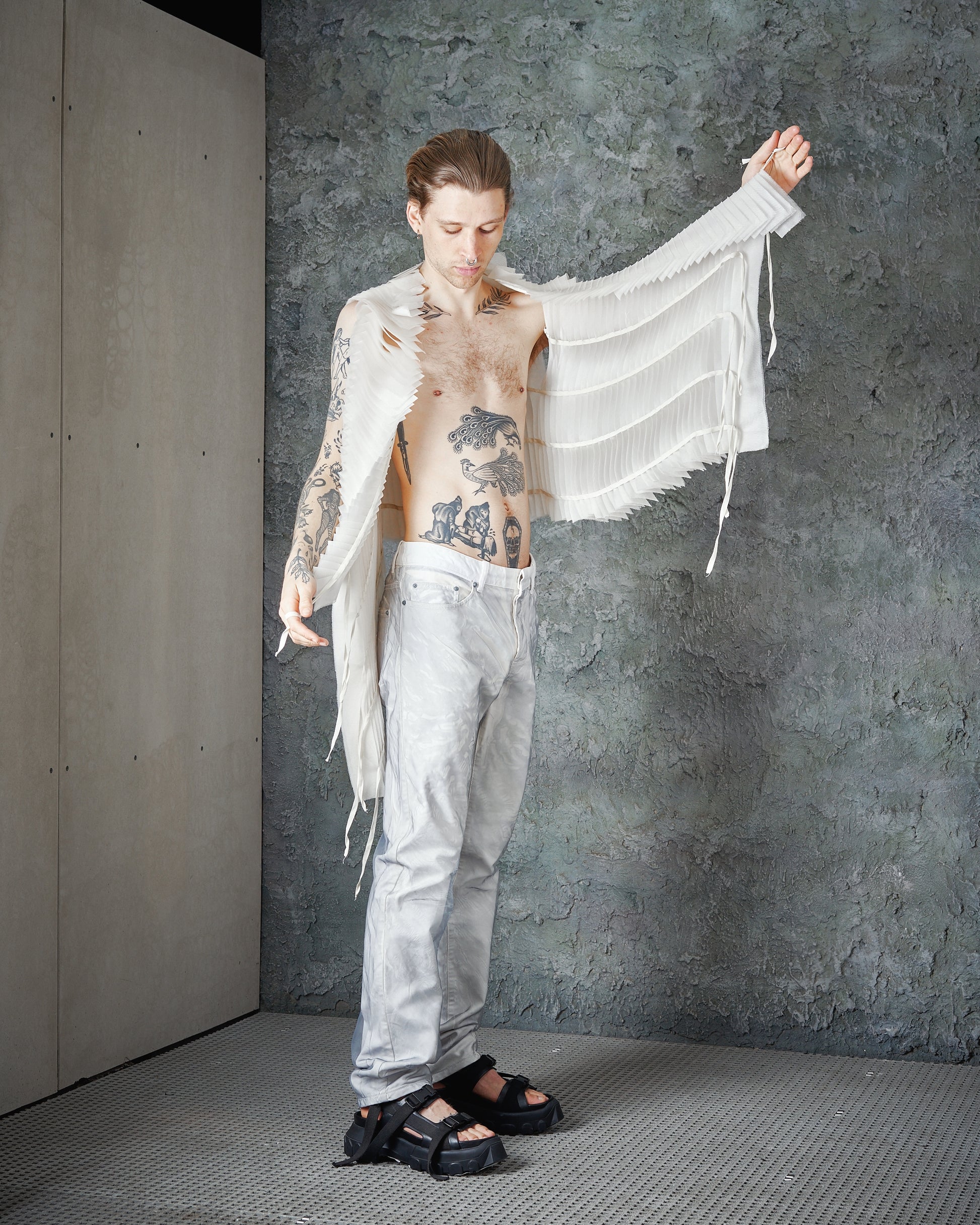 A tattooed person in a textured, flowing white Peachoo + Krejberg top and light pants stands against a rough gray wall, exuding an edgy, artistic vibe.