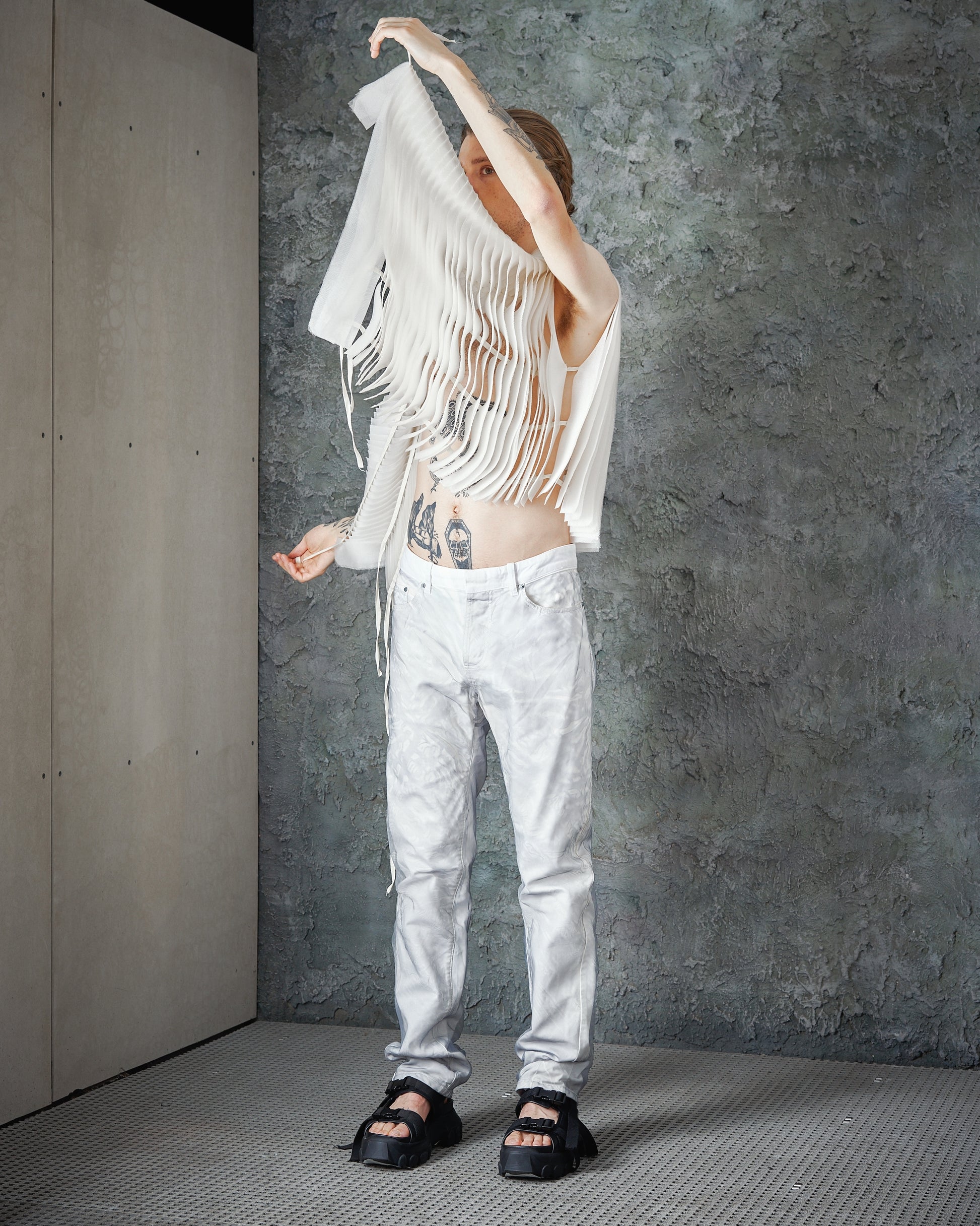Person standing with a raised arm, wearing a fringed, sleeveless white Peachoo + Krejberg top, white pants, and black sandals, set against a textured gray wall.
