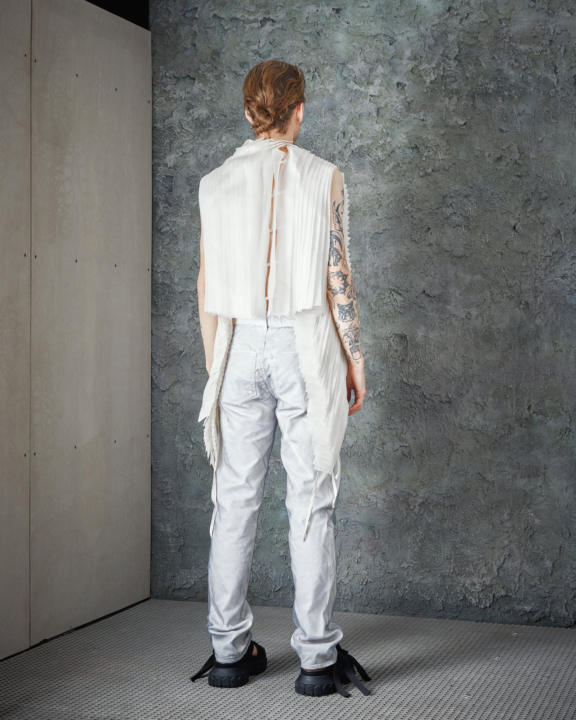 A person with tattoos wears a sleeveless, pleated white Peachoo + Krejberg top and light pants, standing against a textured gray wall. The mood is edgy and artistic.