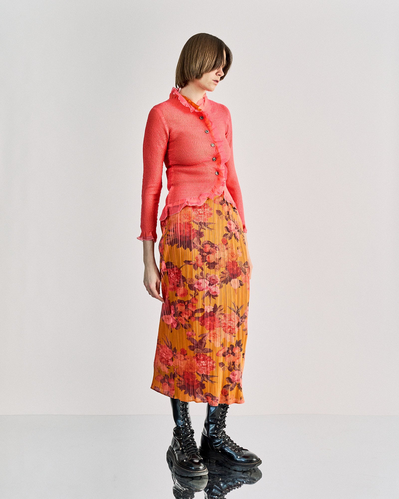 A woman wearing a pink ruffle Peplum by Yoshiki Hishinuma cardigan with black Mother of Pearl buttons and an orange pleated skirt with floral patterns. She is accessorized with a red tie and black boots.