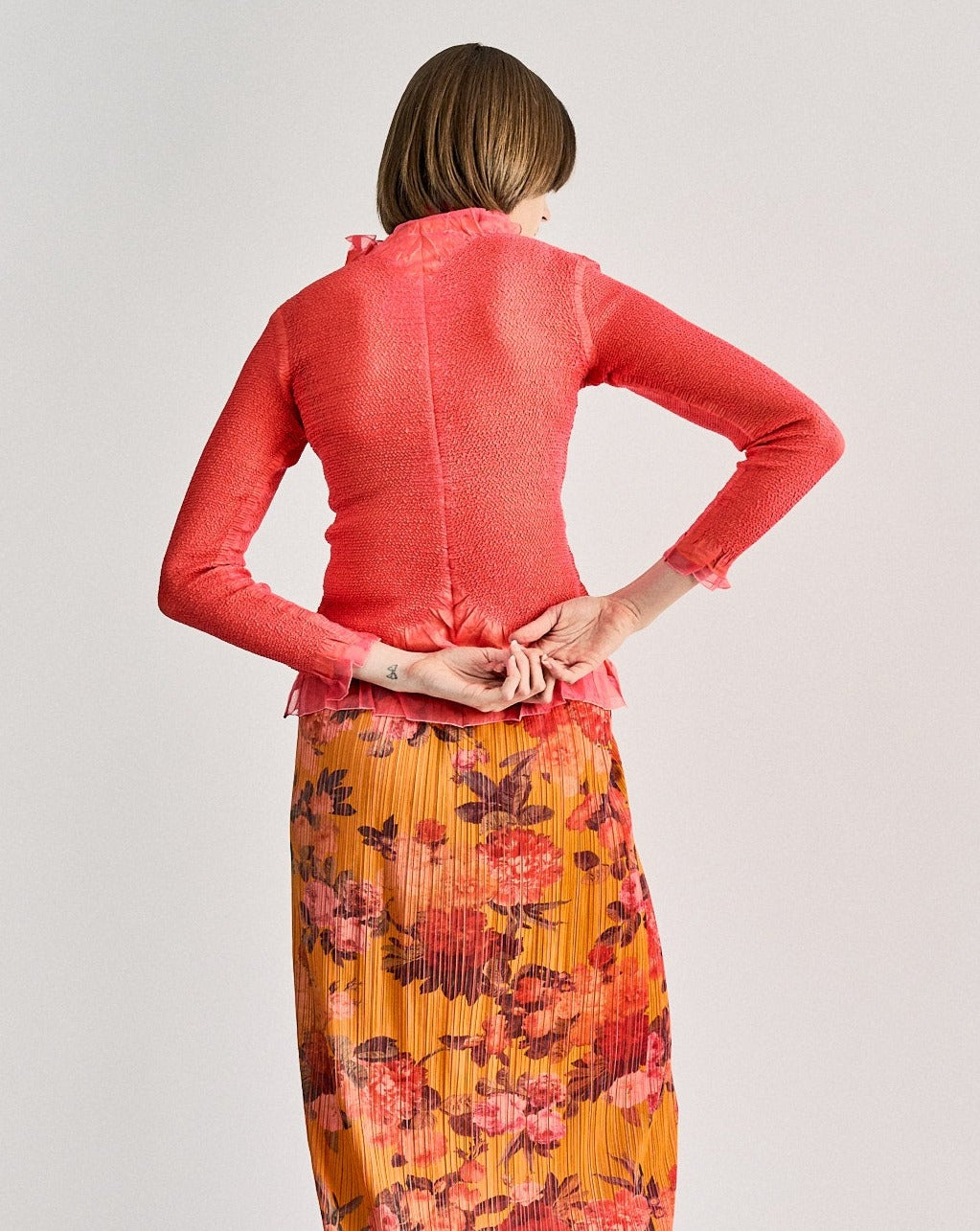 A woman wearing a pink ruffle Peplum by Yoshiki Hishinuma cardigan with black Mother of Pearl buttons and an orange pleated skirt with floral patterns. She is accessorized with a red tie and black boots.