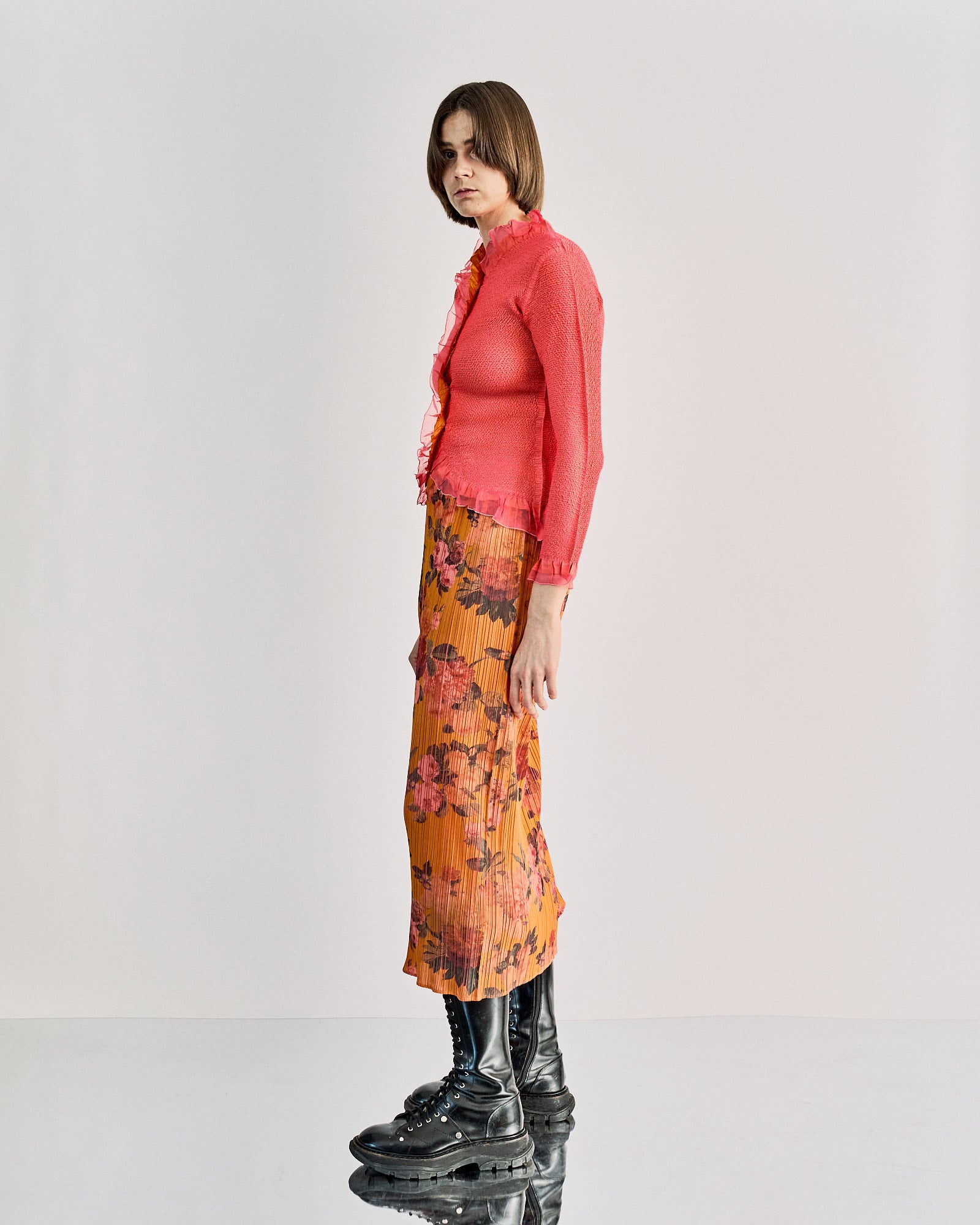 A woman wearing a coral long-sleeve Peplum by Yoshiki Hishinuma top and a pleated orange skirt with pink floral patterns. Her posture is relaxed.