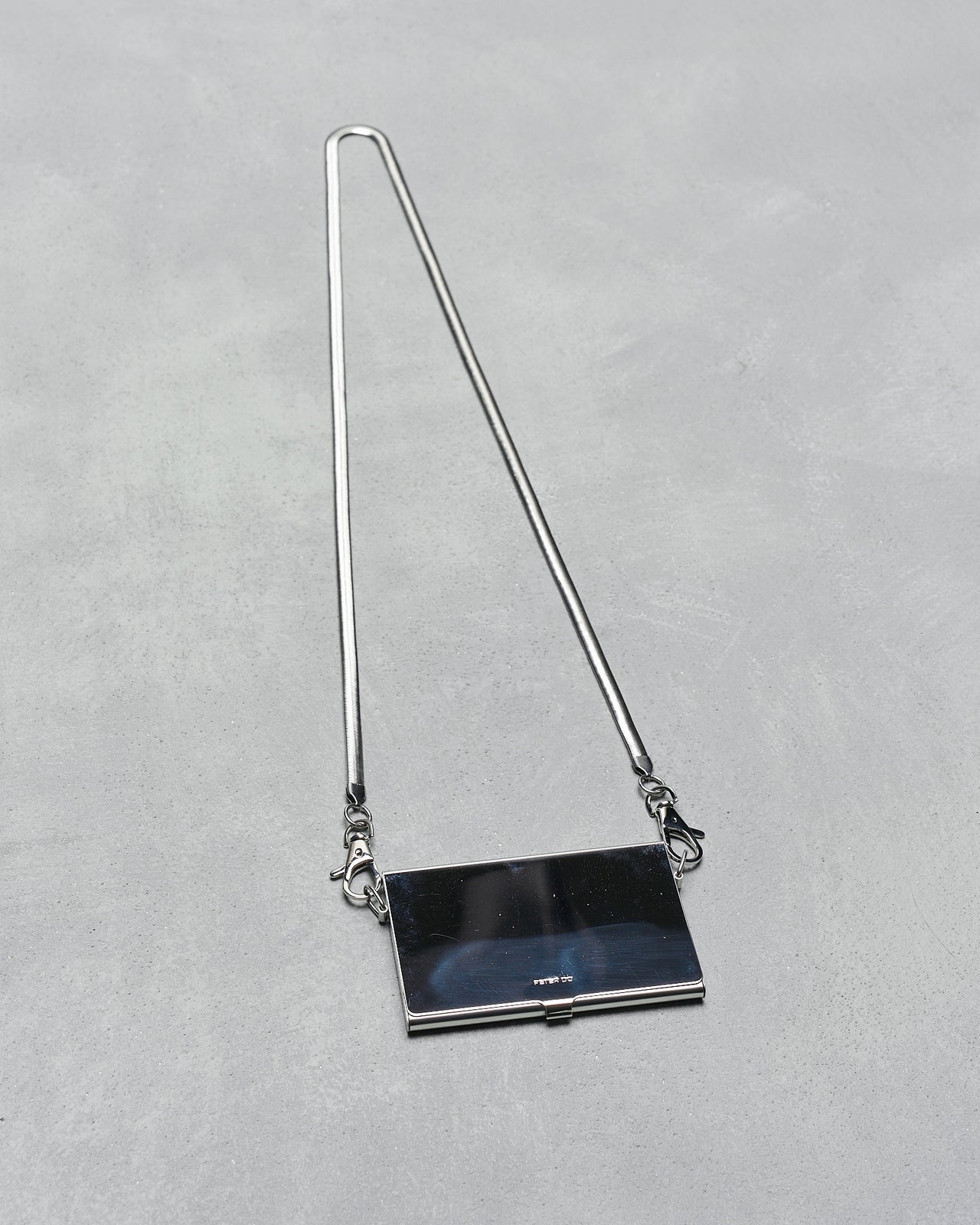 A sleek, rectangular silver clutch from Peter Do with a long strap lies on a textured grey surface. 