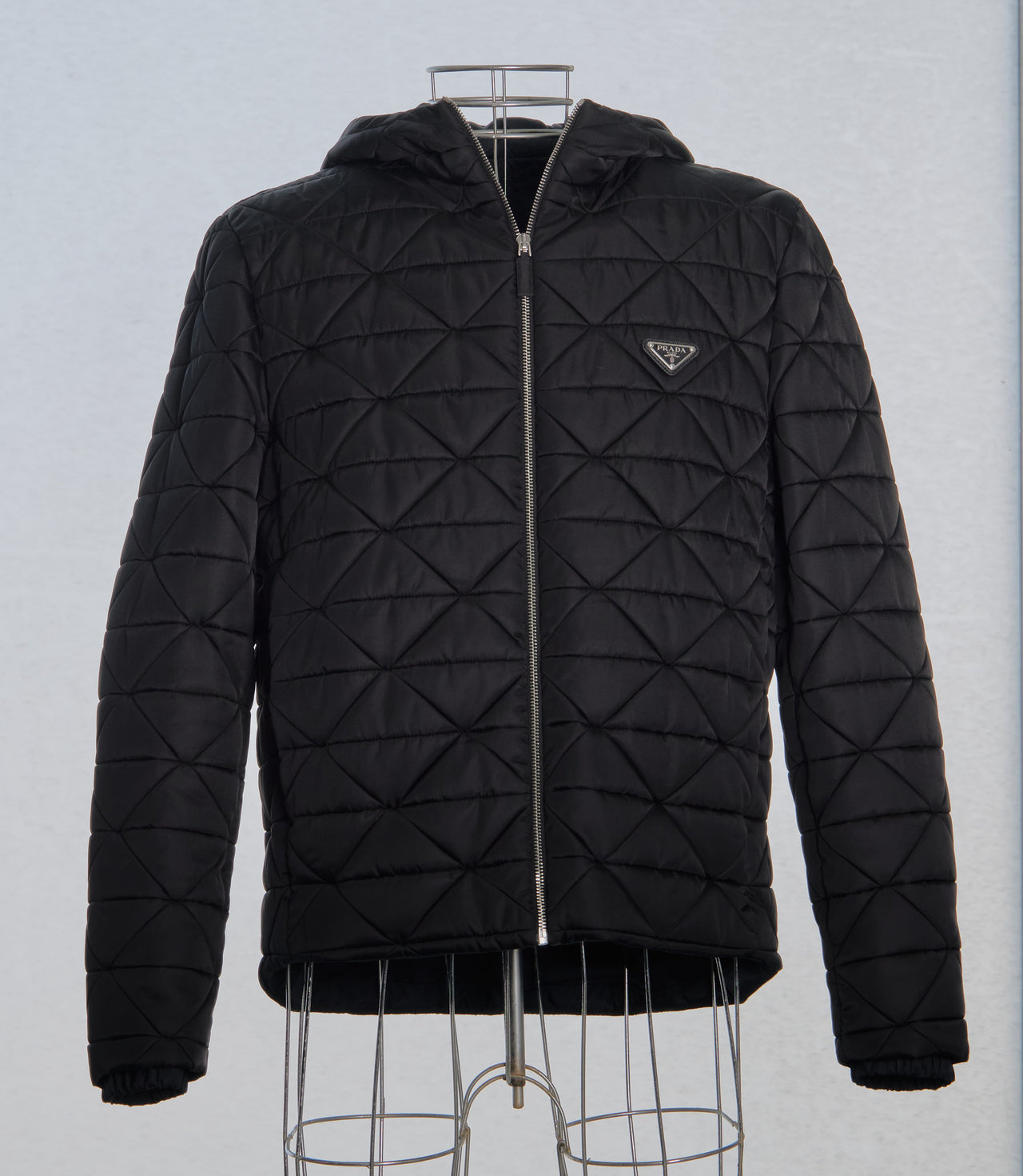 Black quilted Prada jacket with a hood and front zipper, displayed on a wire mannequin. Features a triangular logo patch on the chest, conveying a stylish and modern look.
