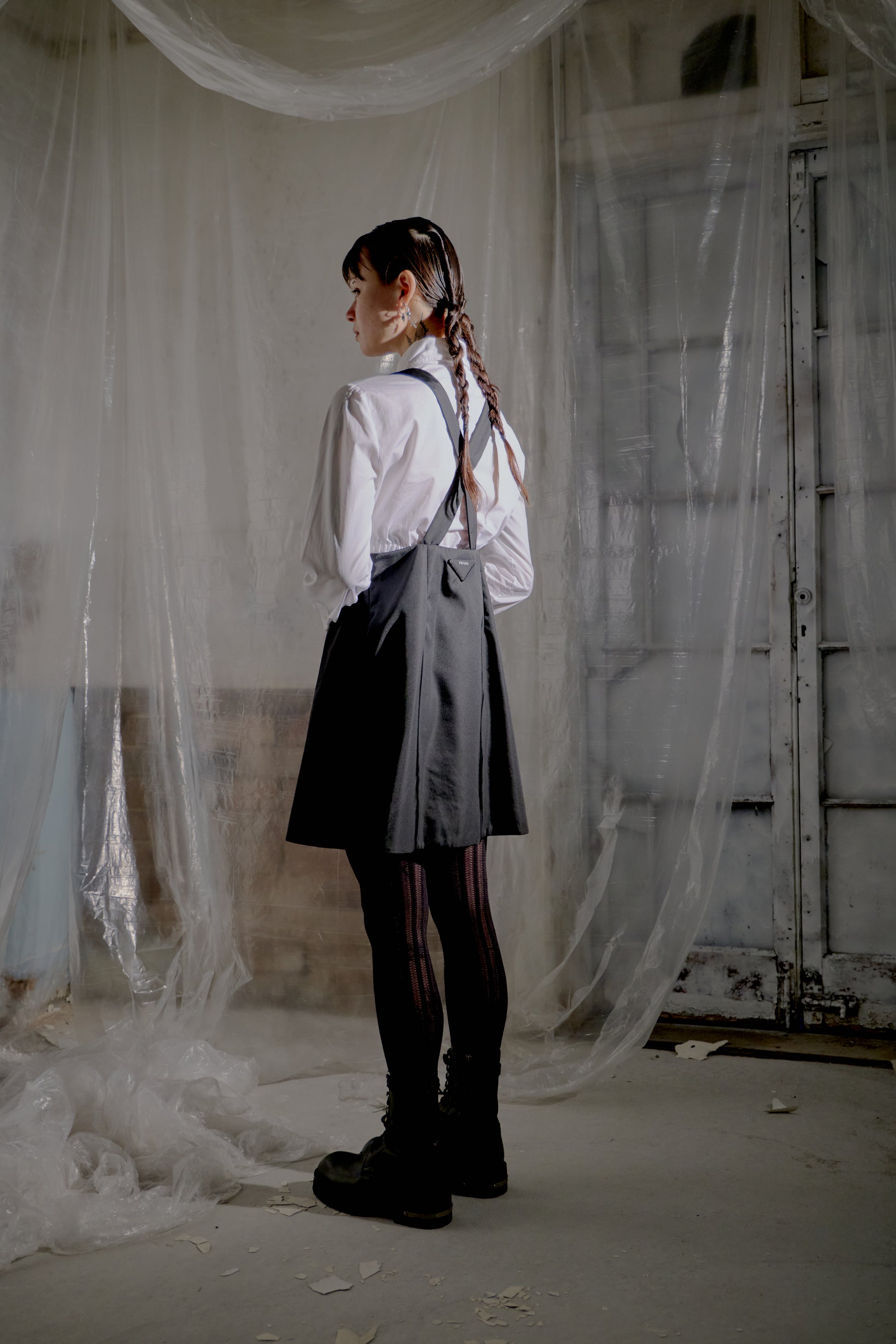 Person wearing a white shirt, gray Prada dress, and black boots in an industrial setting with draped fabric.
